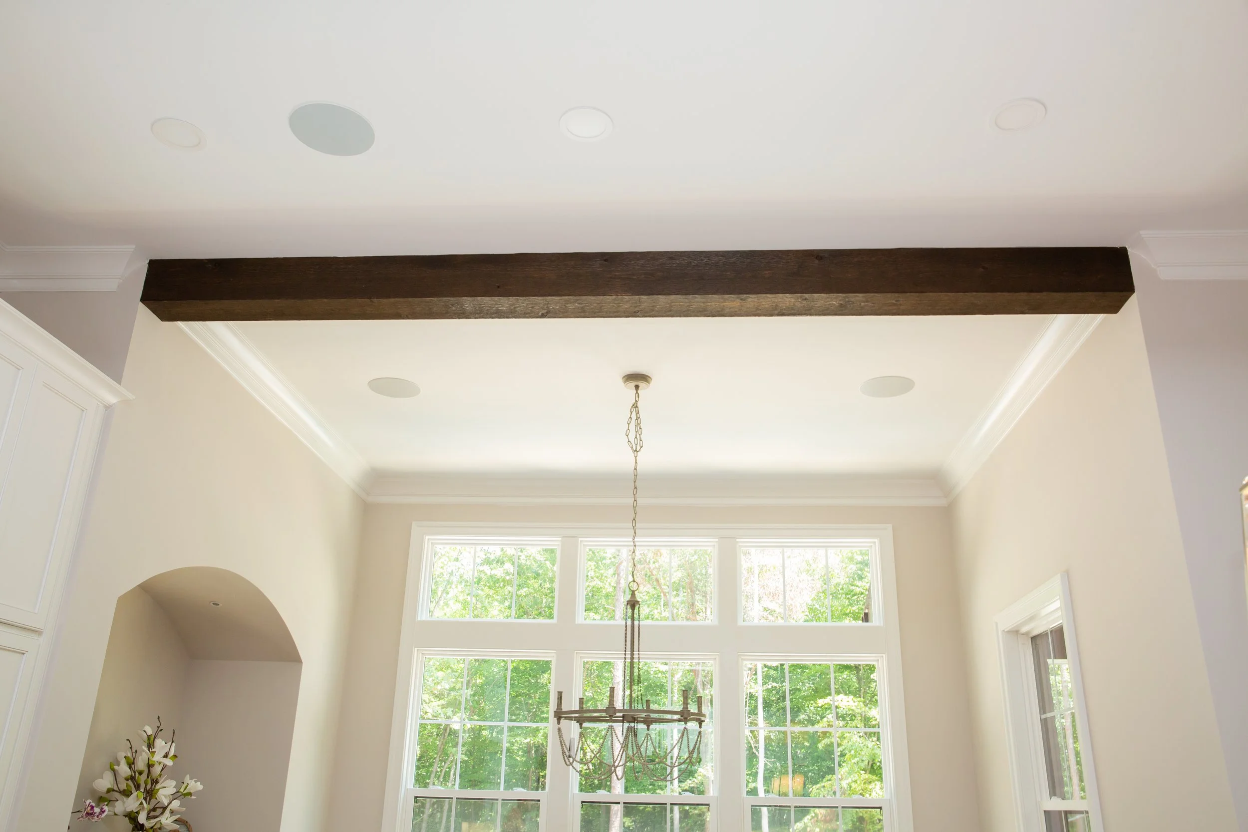 A high ceiling with a large wooden beam and multiple recessed lights, a chandelier hanging from a chain, large windows with lush greenery outside, and part of a decorative floral arrangement in the lower left corner.