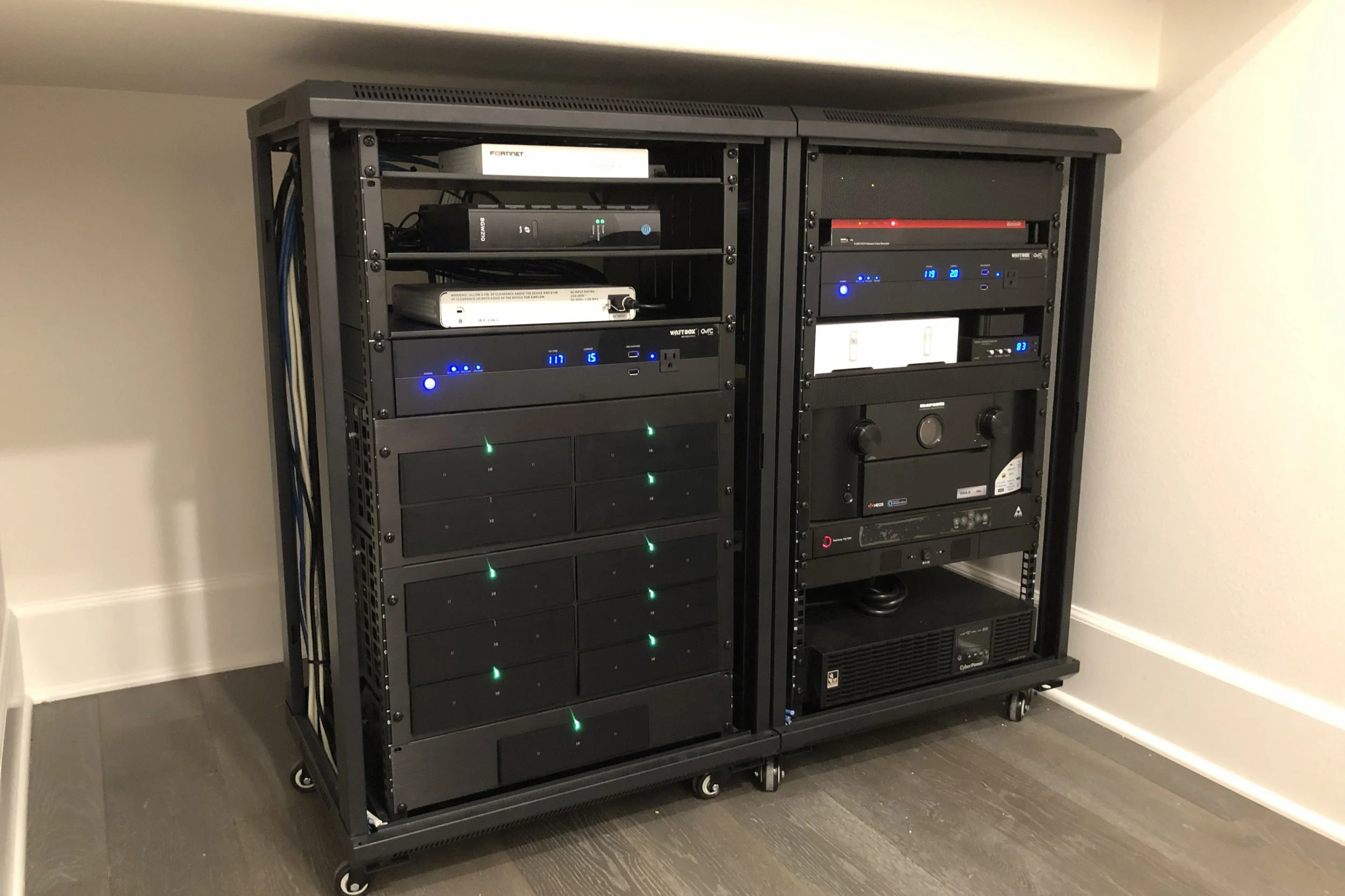 Two black server racks on wheels installed against a beige wall with dark flooring, containing electronic network equipment with blinking lights and cords.