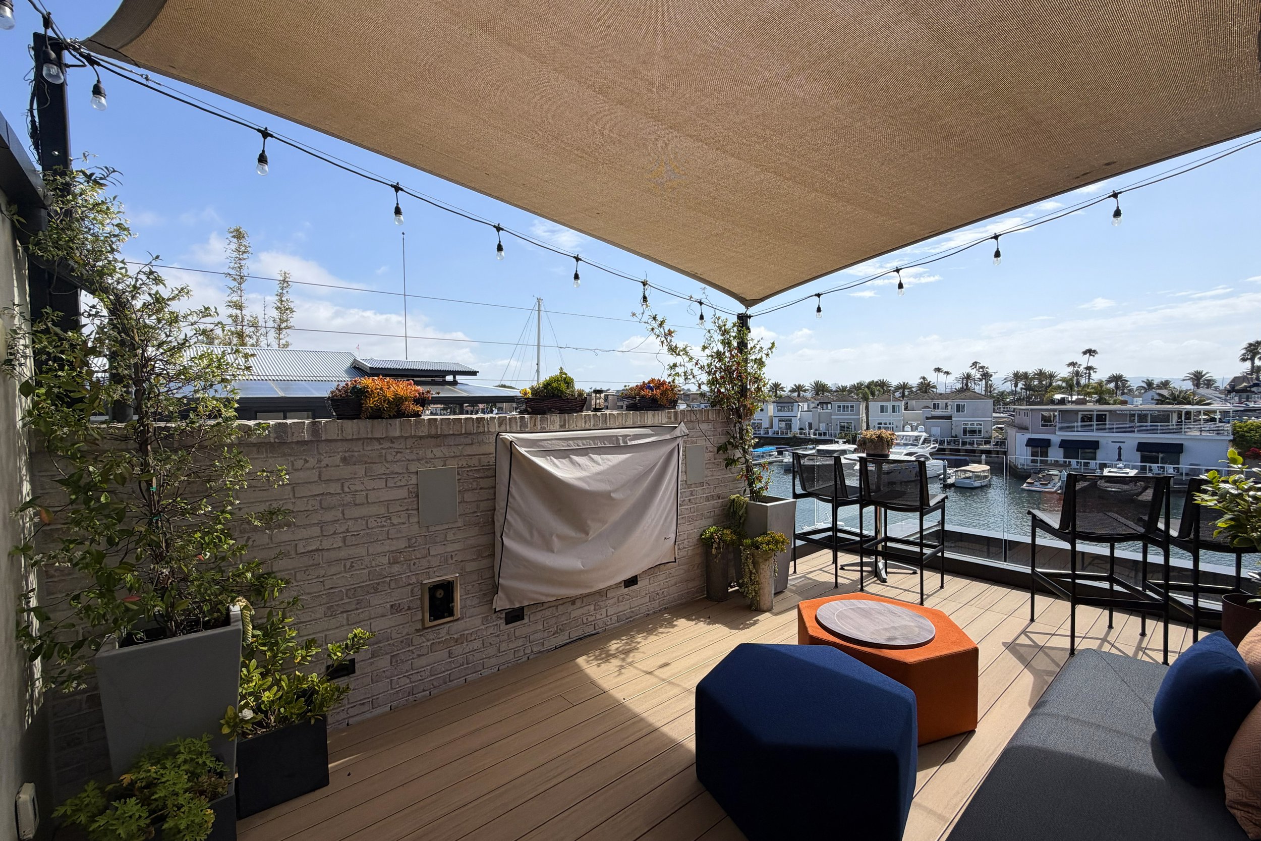 A waterfront outdoor patio or balcony with seating and potted plants, overlooking boats in a marina and a neighborhood of modern houses with palm trees.