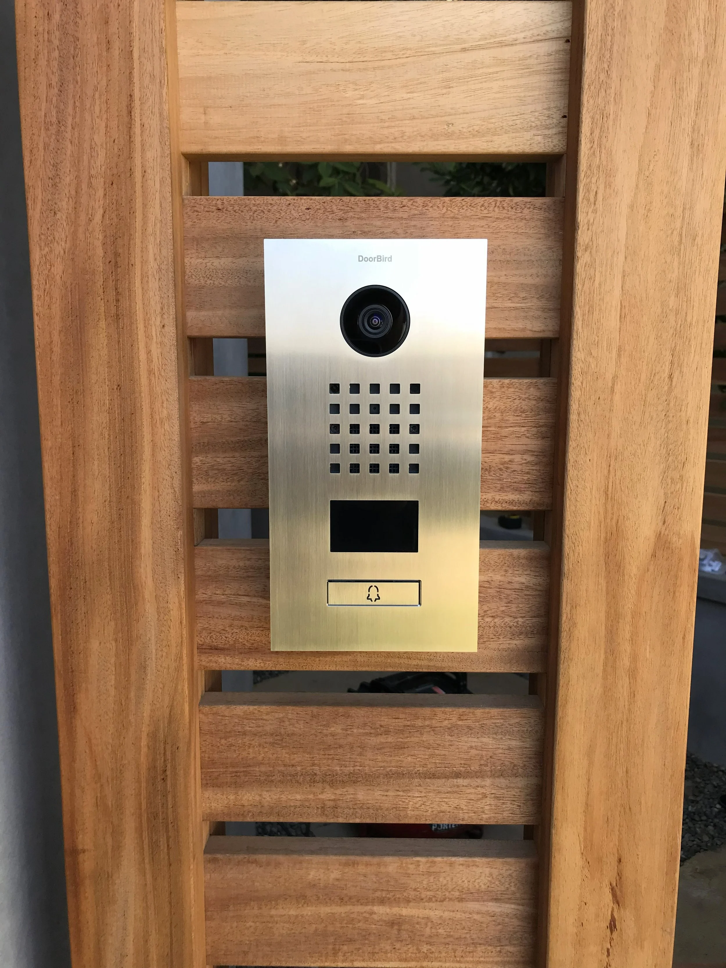 Wooden porch with a metal intercom system featuring a camera, speaker grille, and a doorbell button.