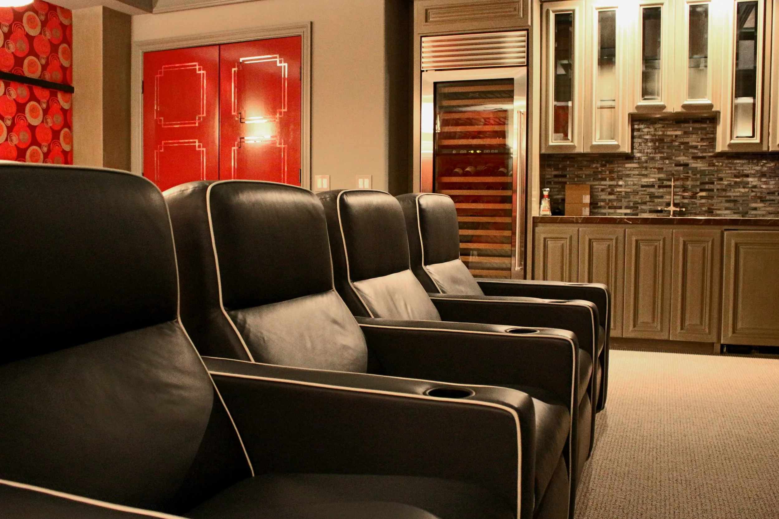 A row of black theater or auditorium seats in front of a kitchenette area with cabinets, a wine cooler, and red patterned wallpaper.