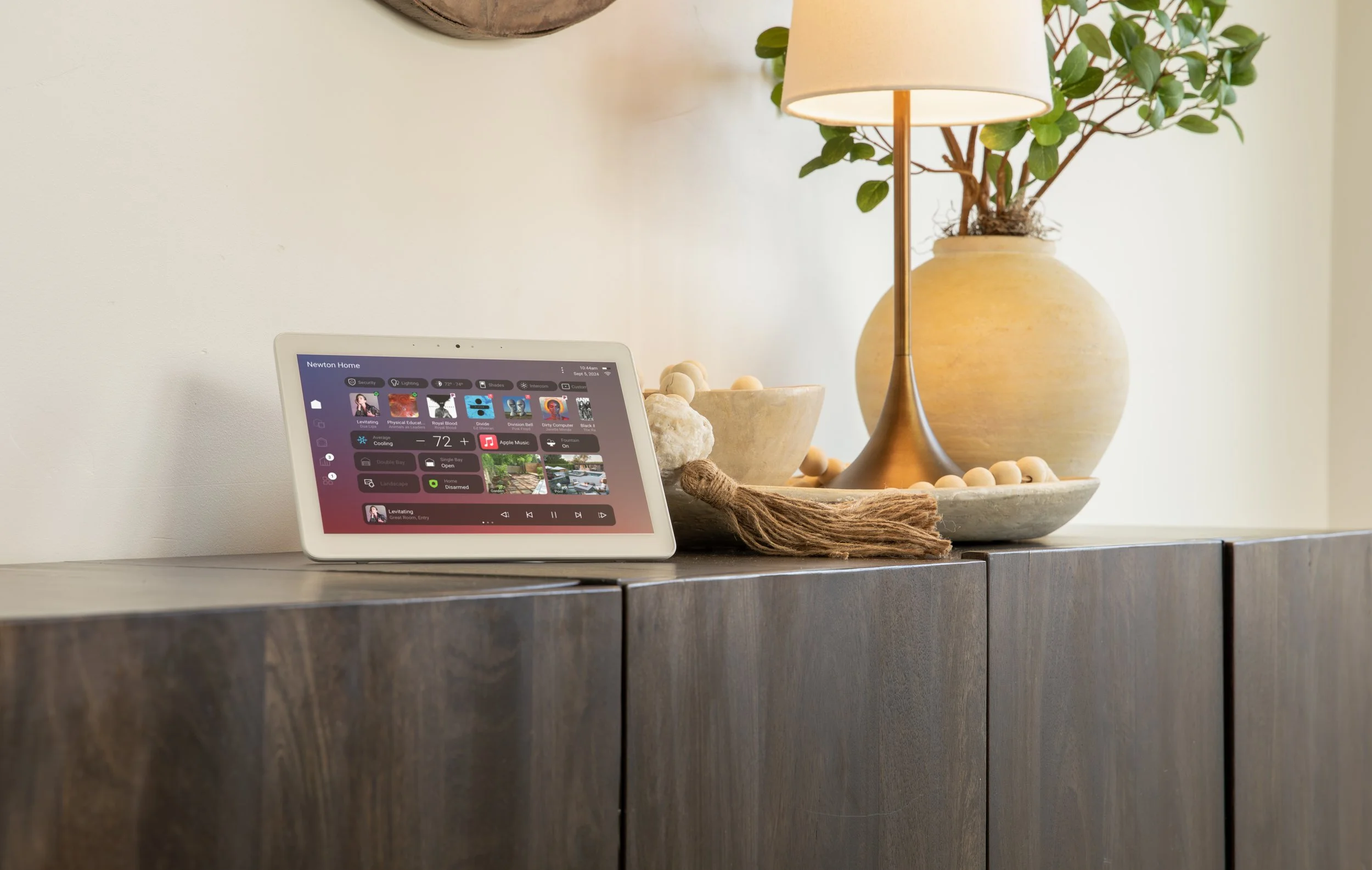 A smart home control device on a wooden cabinet, near decorative vases with plants and a lamp.