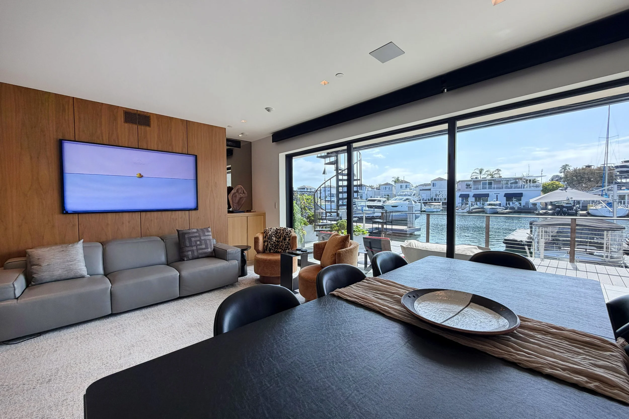 Living room with large windows overlooking a marina with boats, sofa, armchairs, a television, and a dining table.