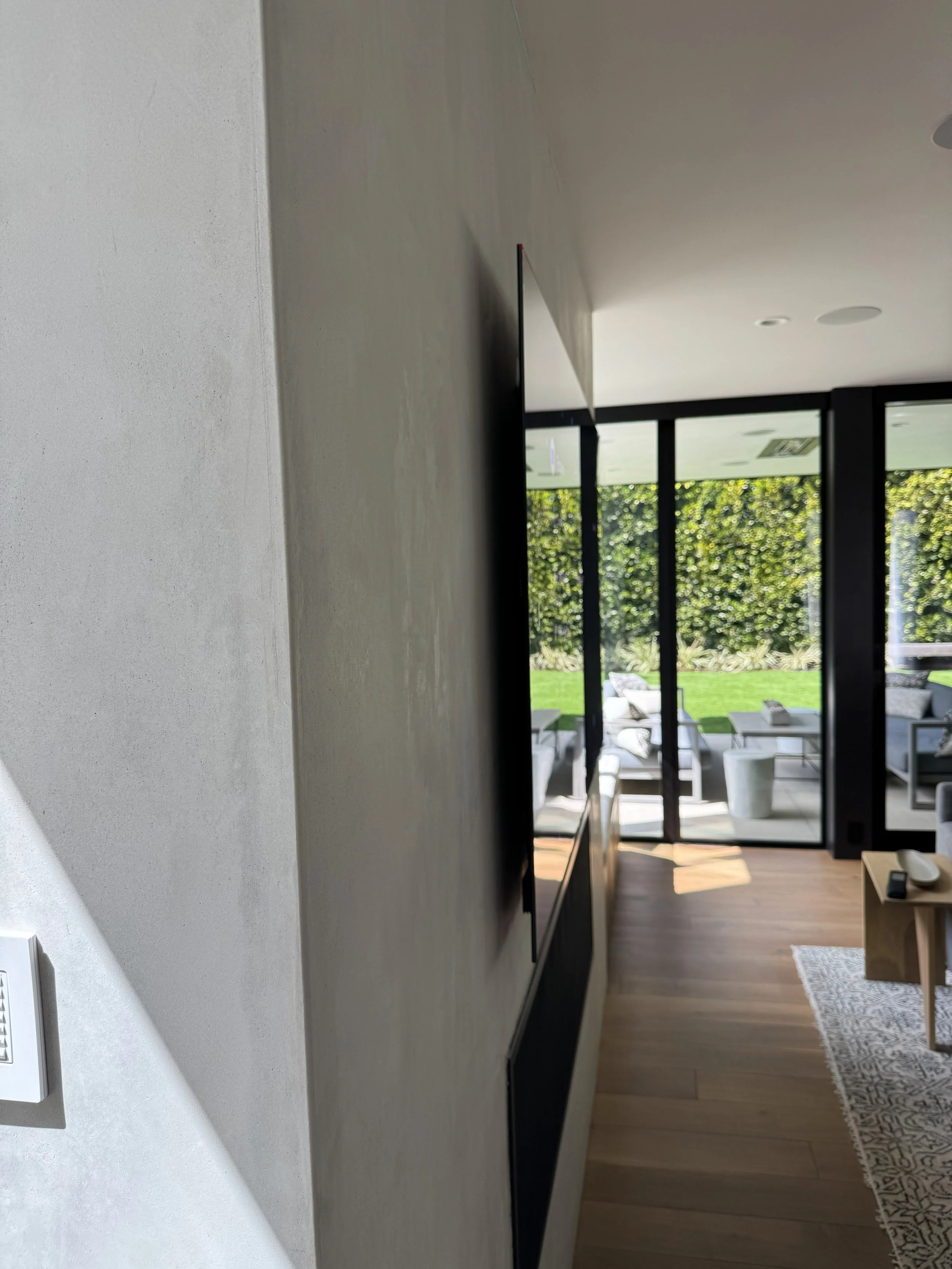 Interior view of a modern living room with large glass doors opening to a garden, with furniture outside visible through the doors.