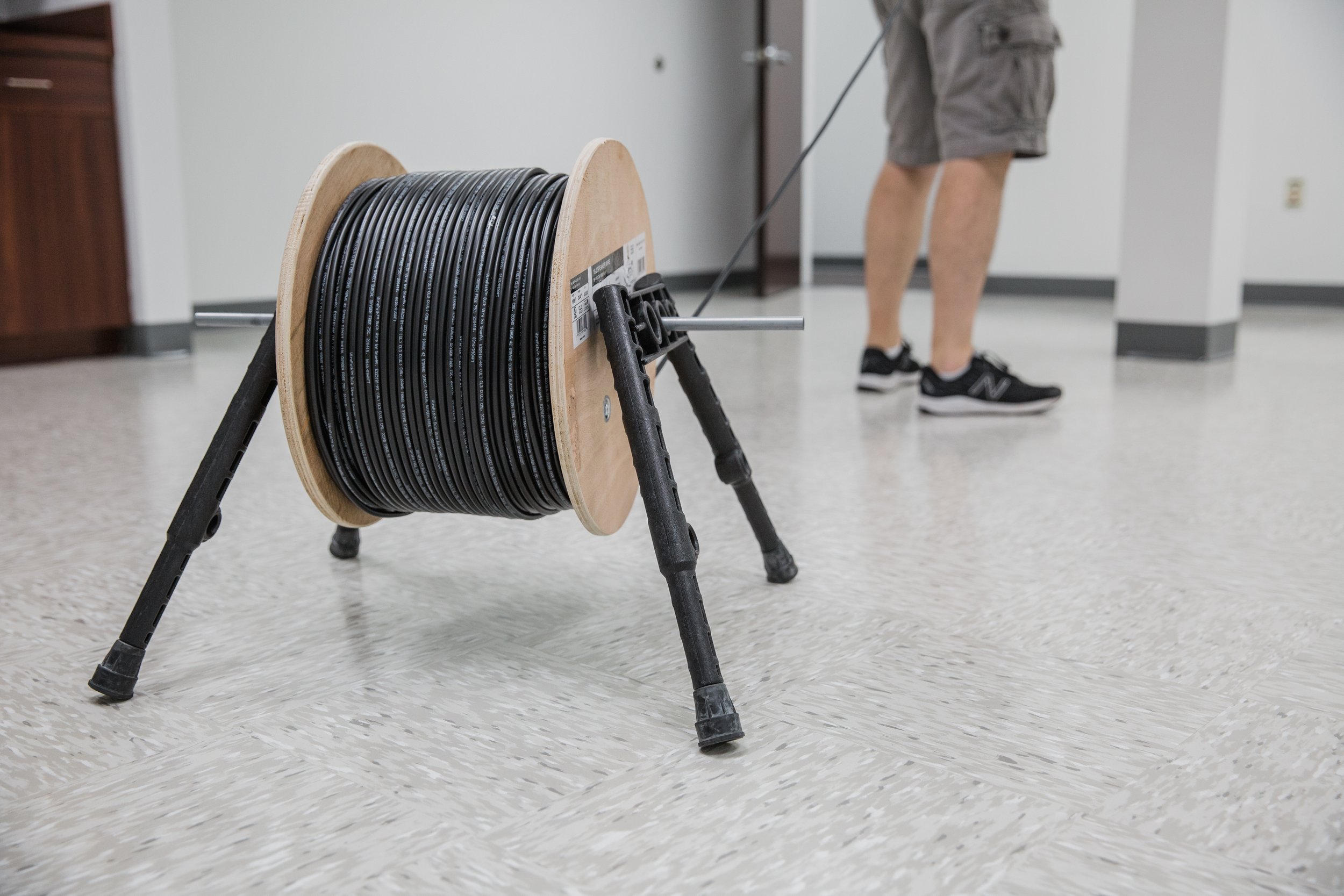A large wooden reel holding several black cables is placed on a metal stand on a light gray tiled floor, with a person in shorts and sneakers visible in the background.