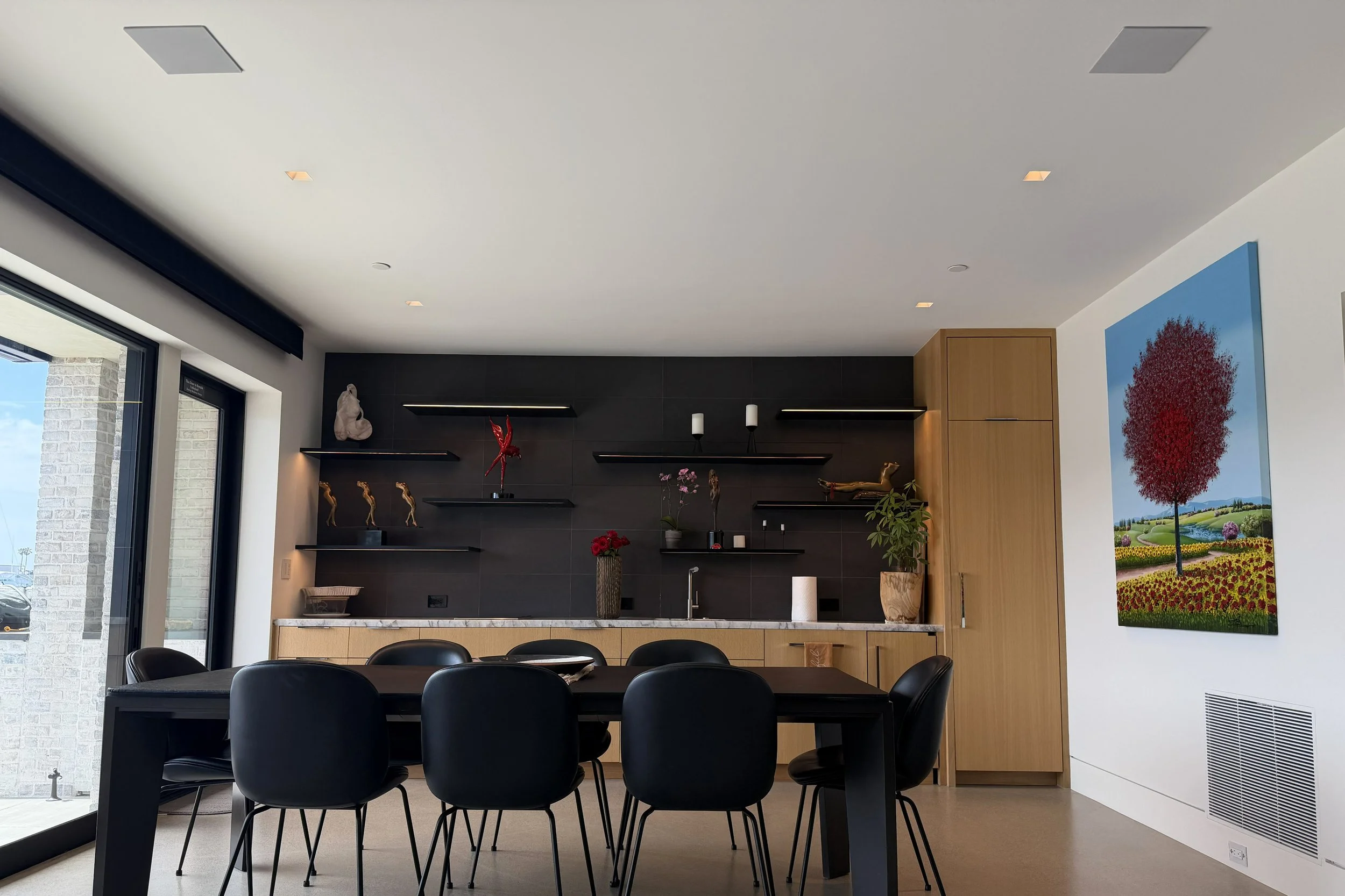 Modern kitchen and dining area with black chairs, a black table, wooden cabinets, a black wall with floating shelves, interior decor, and a large colorful painting of a tree outside the window.