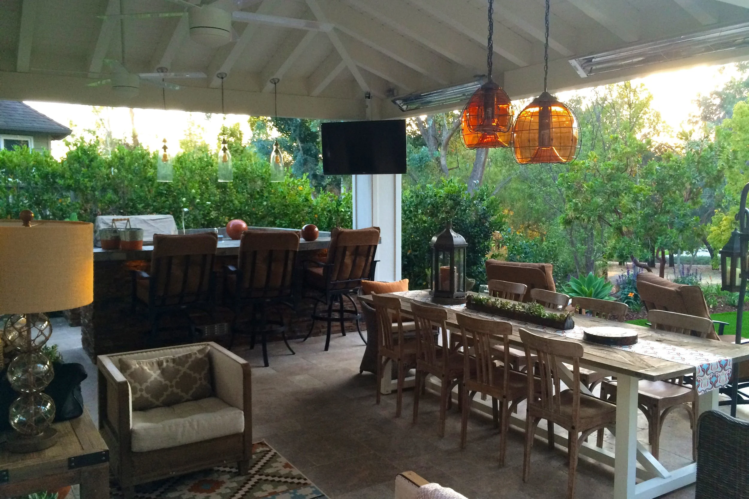 Covered outdoor patio with wooden dining tables and chairs, hanging orange pendant lights, lush greenery, and a black wall-mounted television, illuminated by natural light from the yard and sunset.