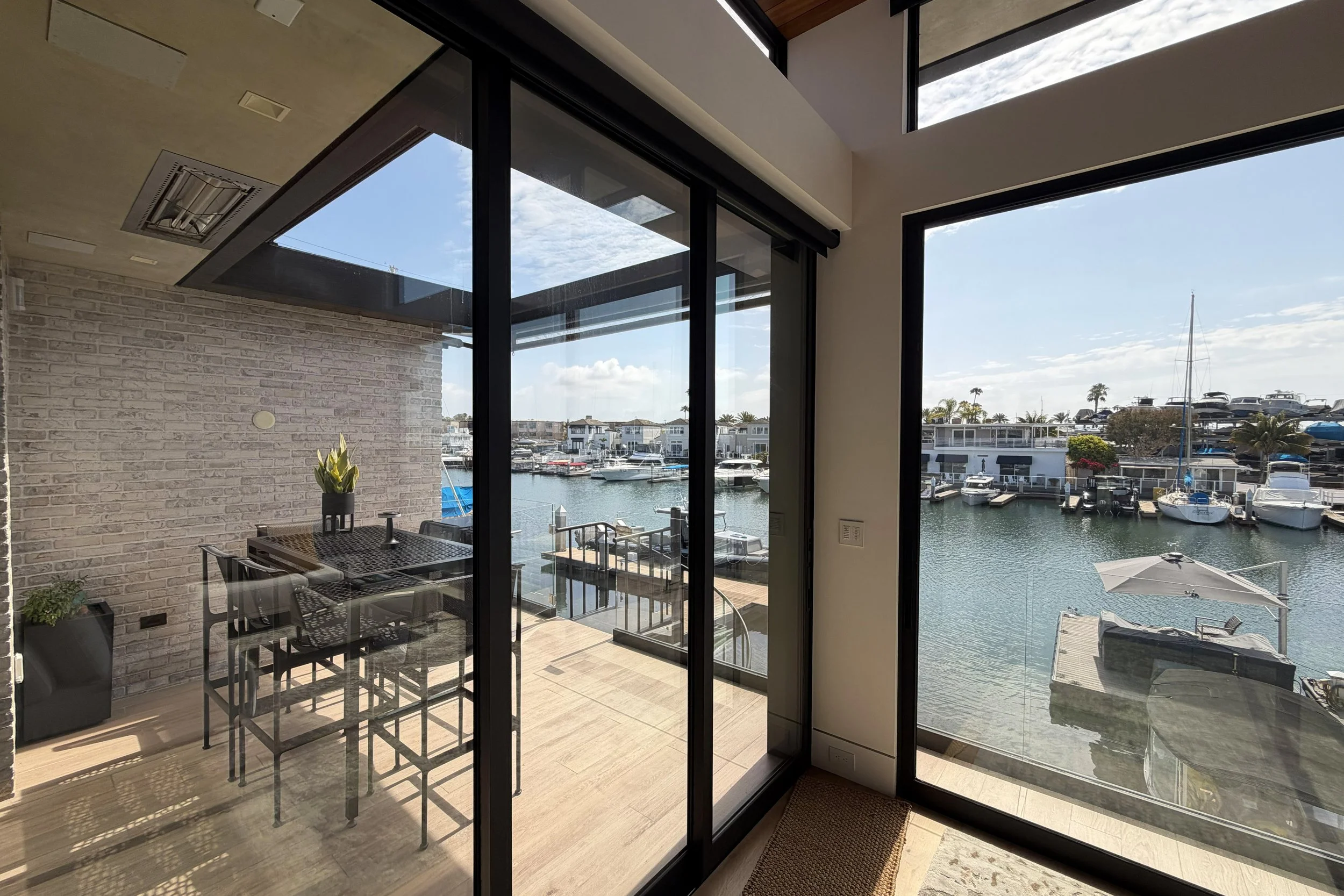Indoor dining area with patio furniture and large glass sliding doors overlooking a marina with boats and houses.