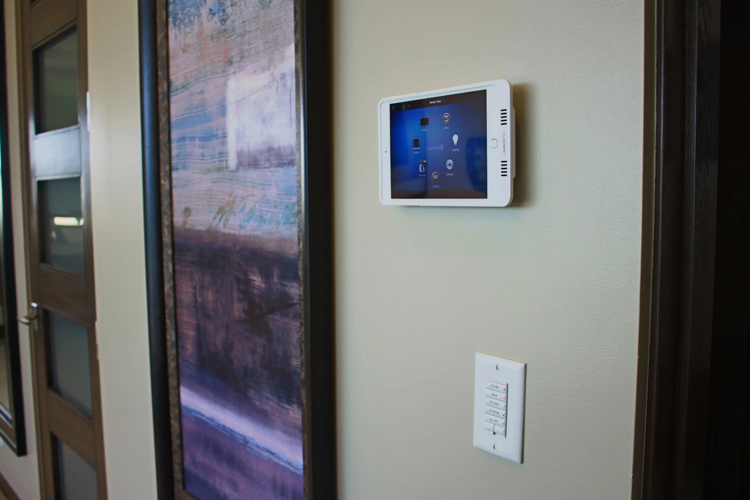 A wall-mounted smart control panel displaying various icons on a blue screen, next to a light switch and a decorative framed mirror or artwork.