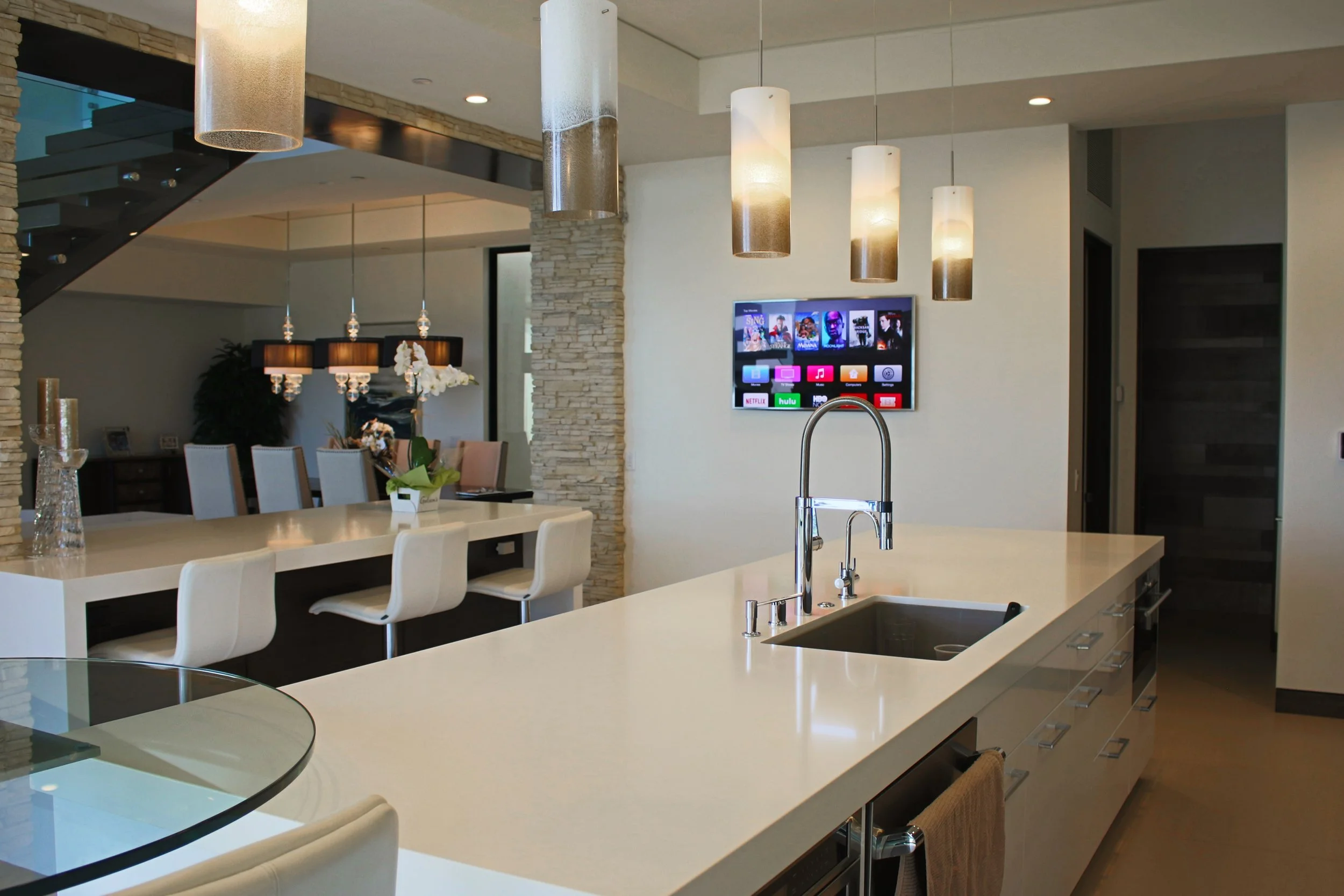 Modern kitchen with white island counter, stainless steel sink, beige chairs, pendant lighting, stone accent wall, flat-screen TV, and dining area in background.