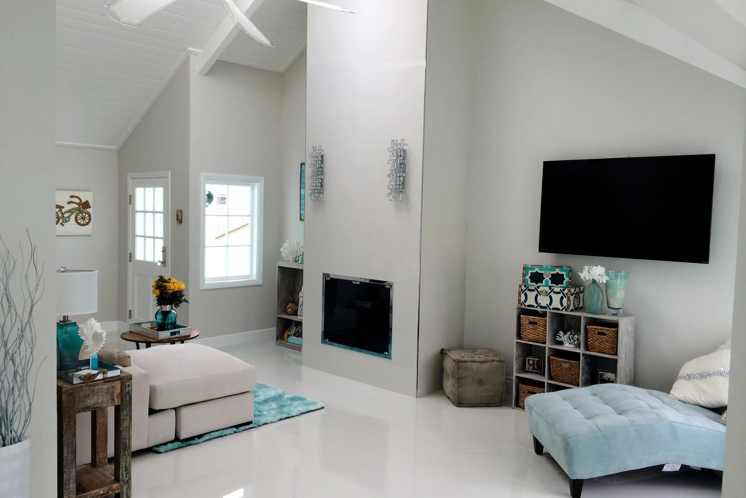 Living room with white walls and ceiling, featuring a large flat-screen TV on the wall, a beige sectional sofa, a tufted ottoman, a small round coffee table with a flower vase, and a bookshelf with decorative items and baskets. There are two windows 