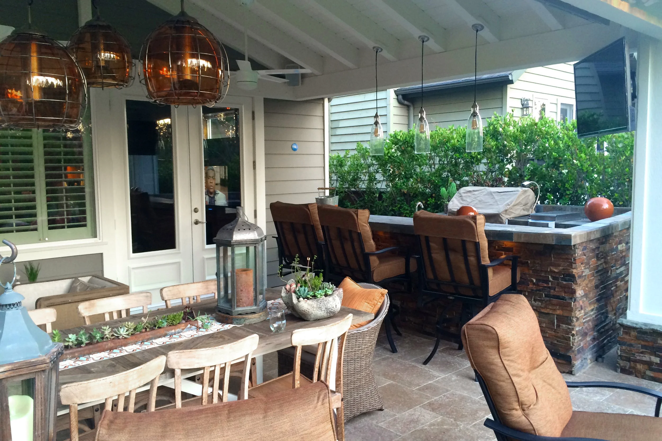 A covered outdoor patio with wooden dining table and chairs, a brick fireplace with a grill, decorative lamps, a TV, and lush greenery in the background.