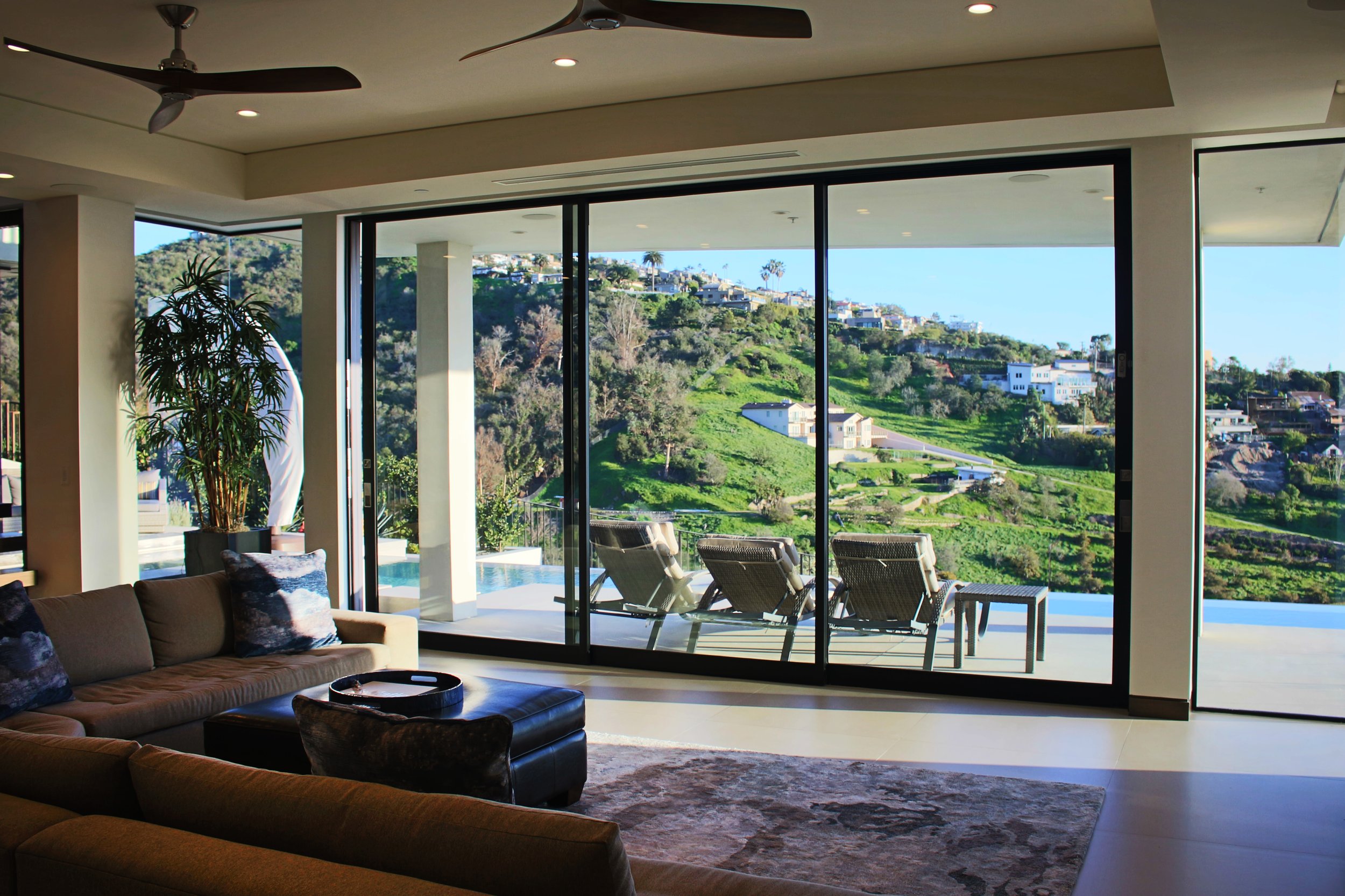 Modern living room with large glass sliding doors overlooking a hillside with houses and greenery. Inside, there is a brown sectional sofa with decorative pillows, a black ottoman with a tray, a potted plant, and a ceiling fan.