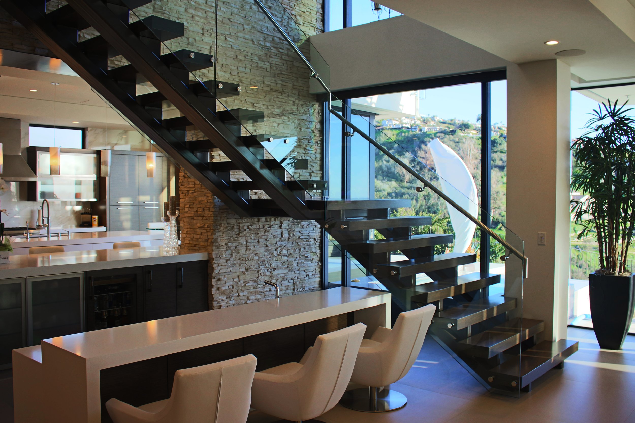 Modern interior of a house featuring a floating staircase with glass railings, stone accent wall, large windows with scenic views, white dining chairs around a table, and indoor plants.