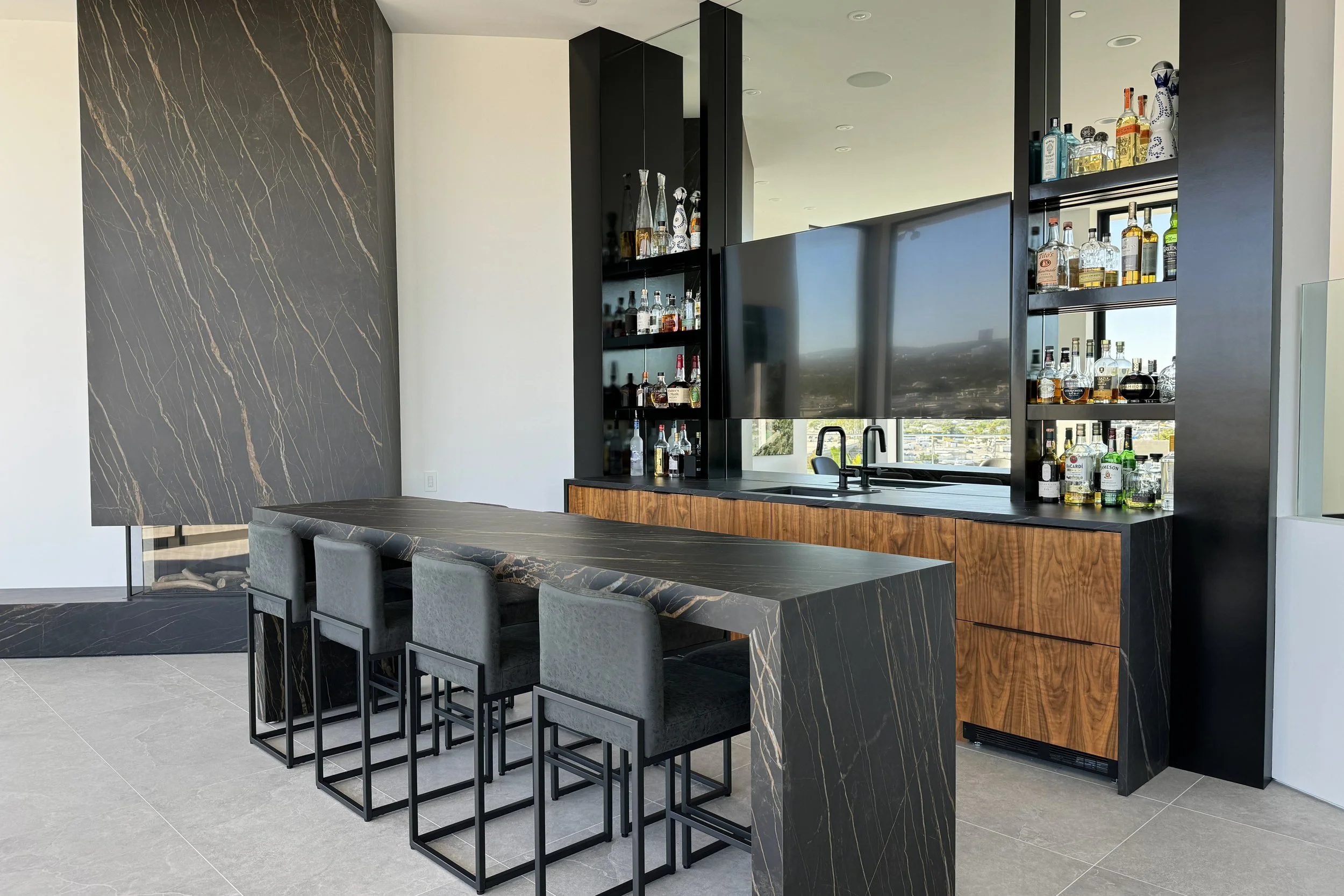 Modern kitchen with black marble island and bar area, gray bar stools, dark wood accents, black shelving with liquor bottles, and large window view.