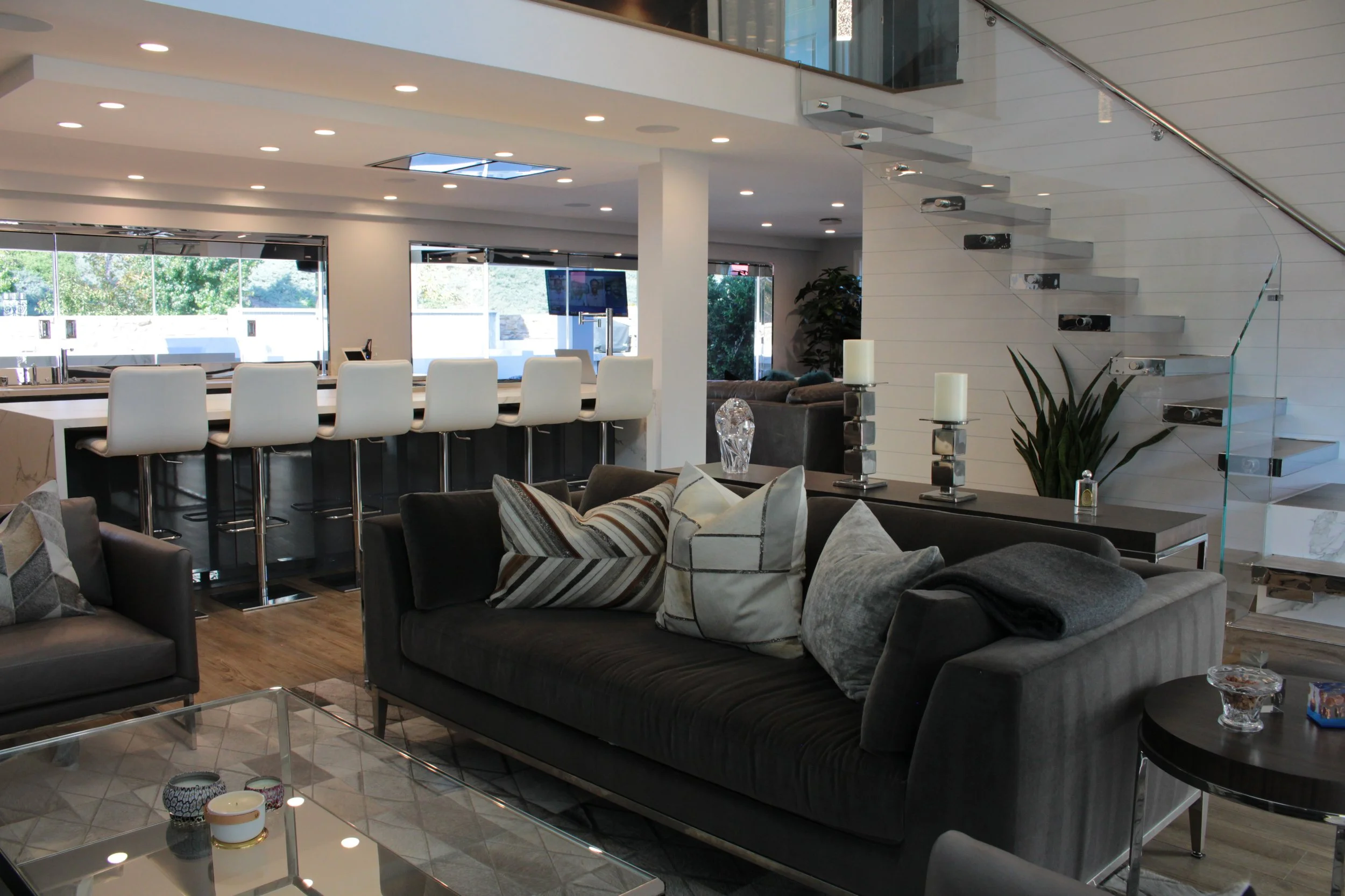 Interior of a modern, brightly lit living and dining area featuring a dark sofa with patterned pillows, a glass coffee table, and a white kitchen island with white bar stools. Large windows and a staircase with glass panels are also visible.