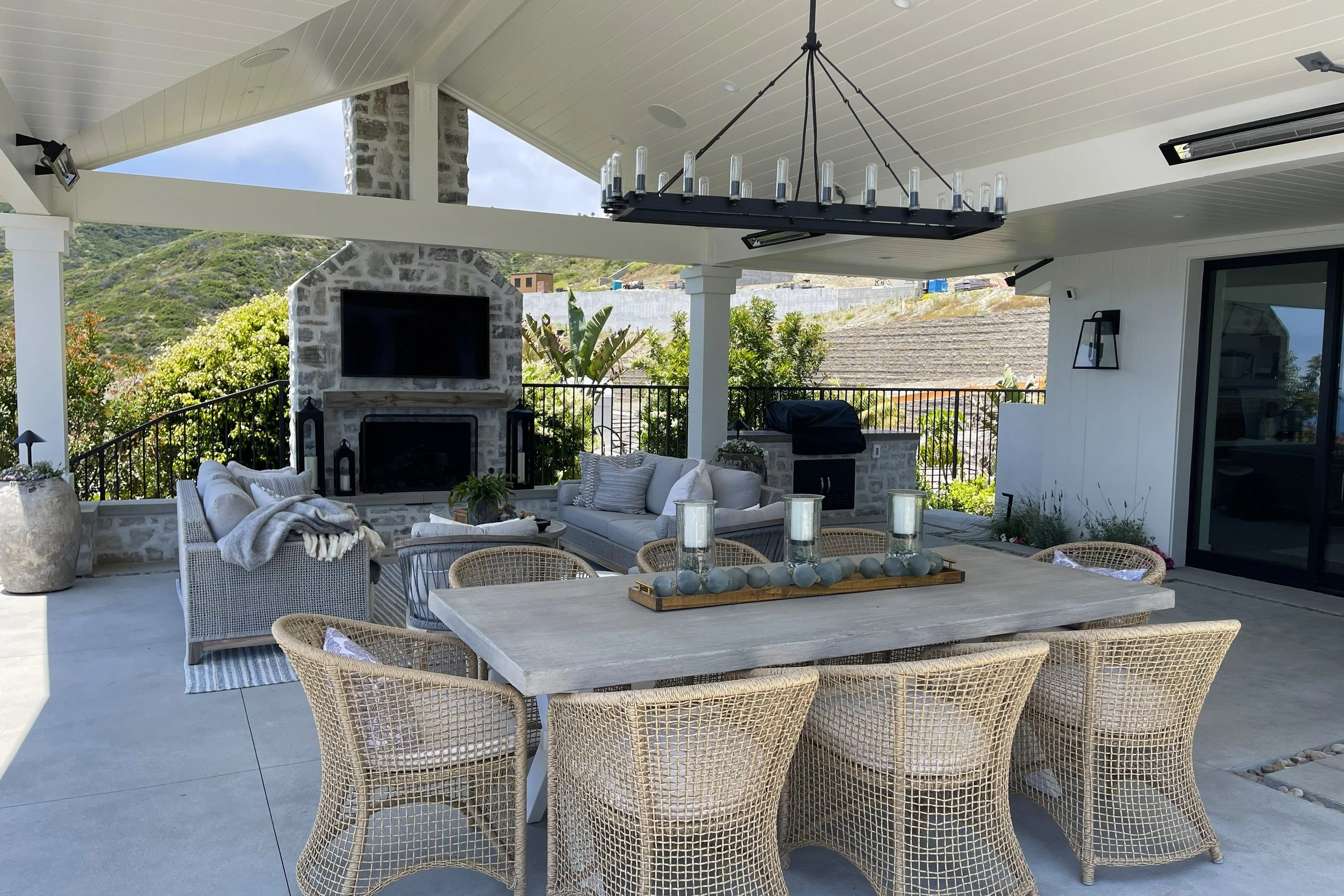Outdoor patio with wicker chairs around a table, a couch, a fireplace with a television, greenery in the background, and a modern chandelier hanging from the ceiling.