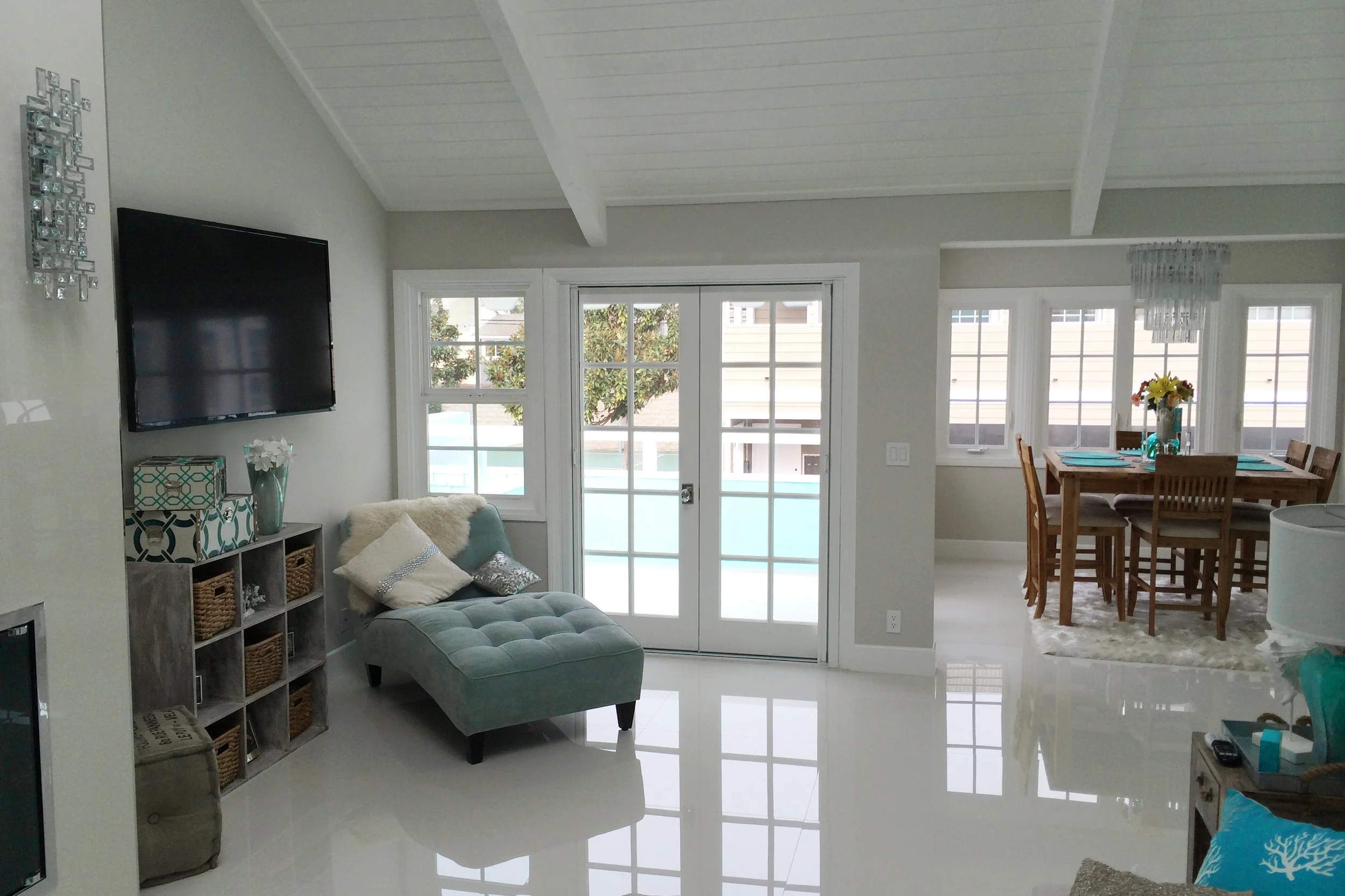 Living room with large windows and glass doors, a TV mounted on the wall, a teal tufted ottoman, a sectional sofa with pillows, a decorative shelving unit with baskets, and a nearby dining area with a wooden table and chairs.
