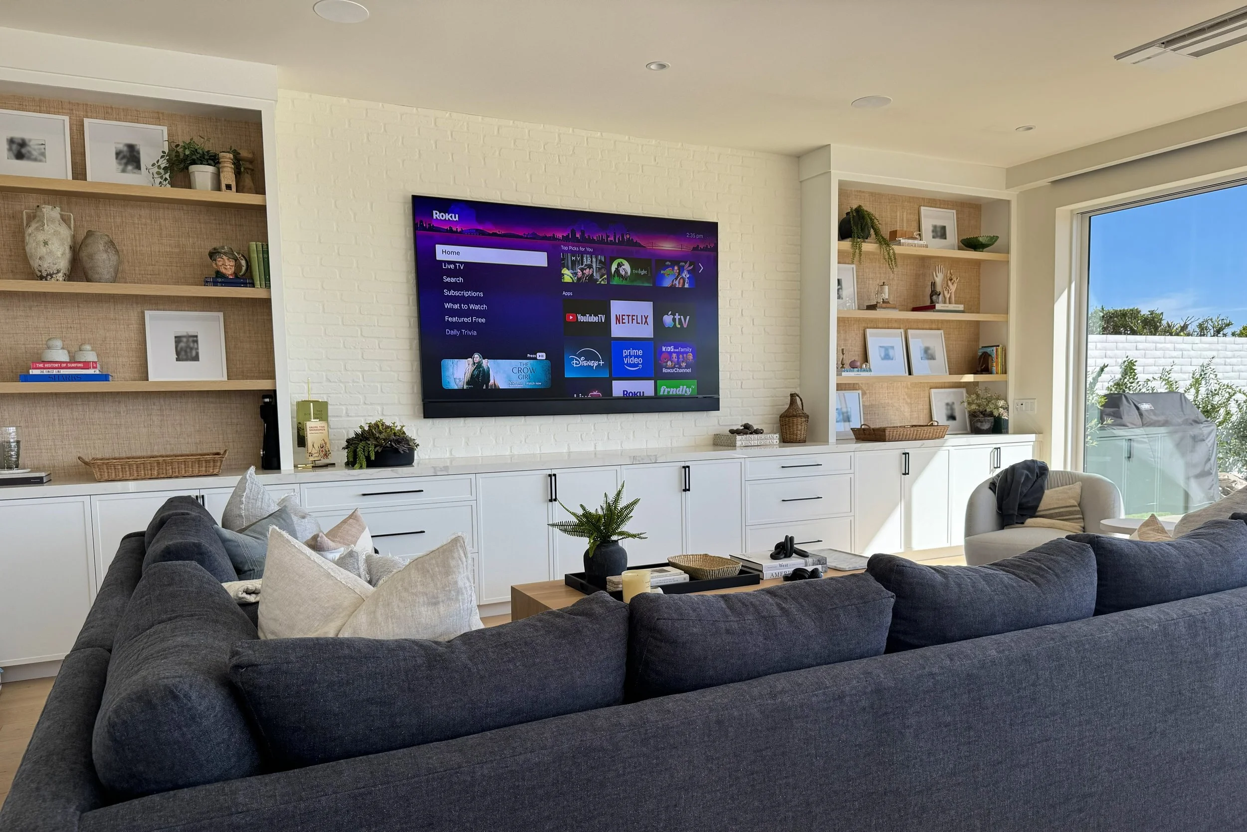 Living room with a large gray sectional sofa, decorative pillows, a wooden coffee table with candles, and built-in white cabinets with open shelving filled with artwork, vases, books, and plants. A large flat-screen TV is mounted on a white brick wal