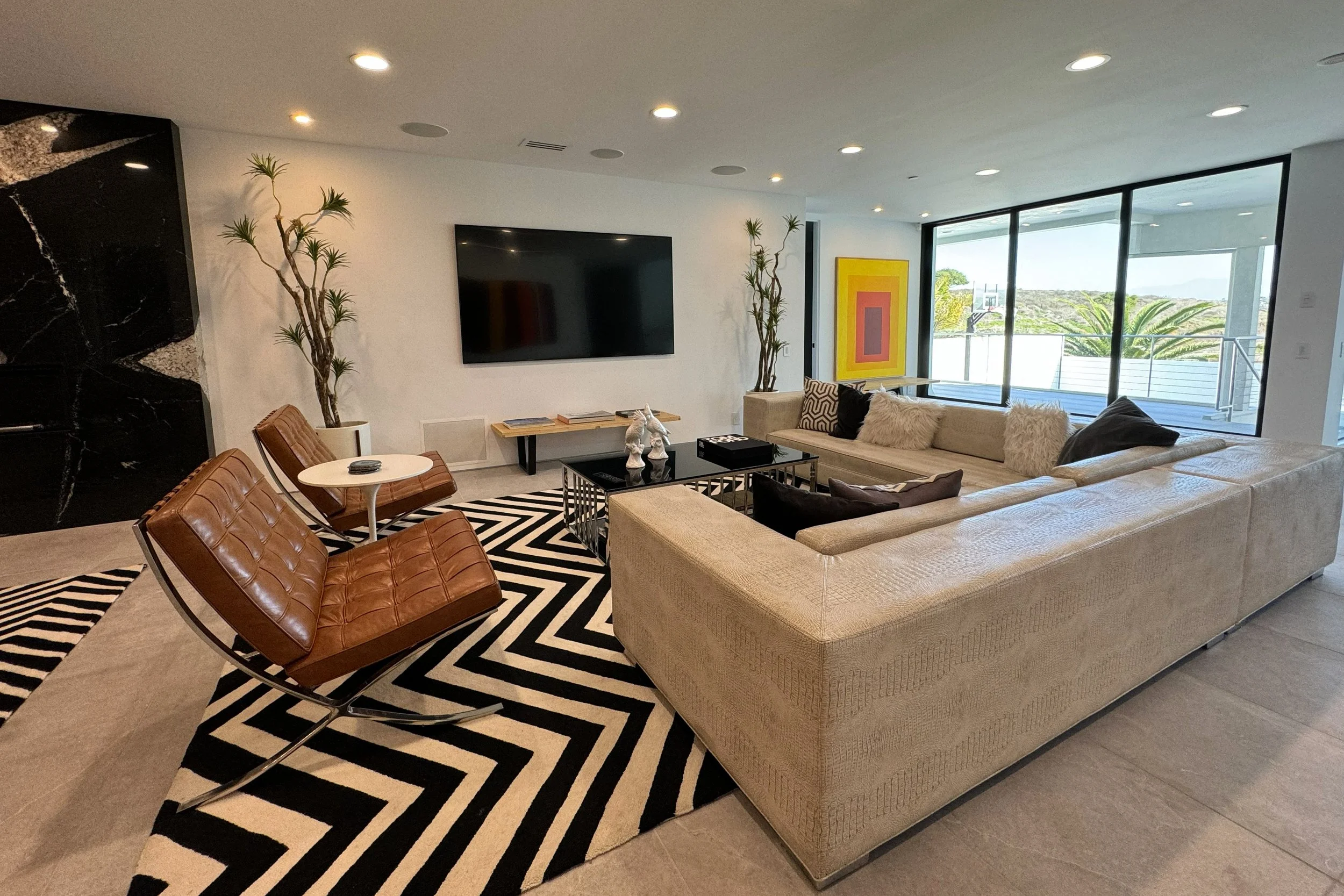 Modern living room with a beige sectional sofa with fluffy pillows, a glass coffee table,  two retro-style leather chairs, a large flat-screen TV on the wall, decorative plants, artwork on the wall, and large windows with a view of palm trees and a b