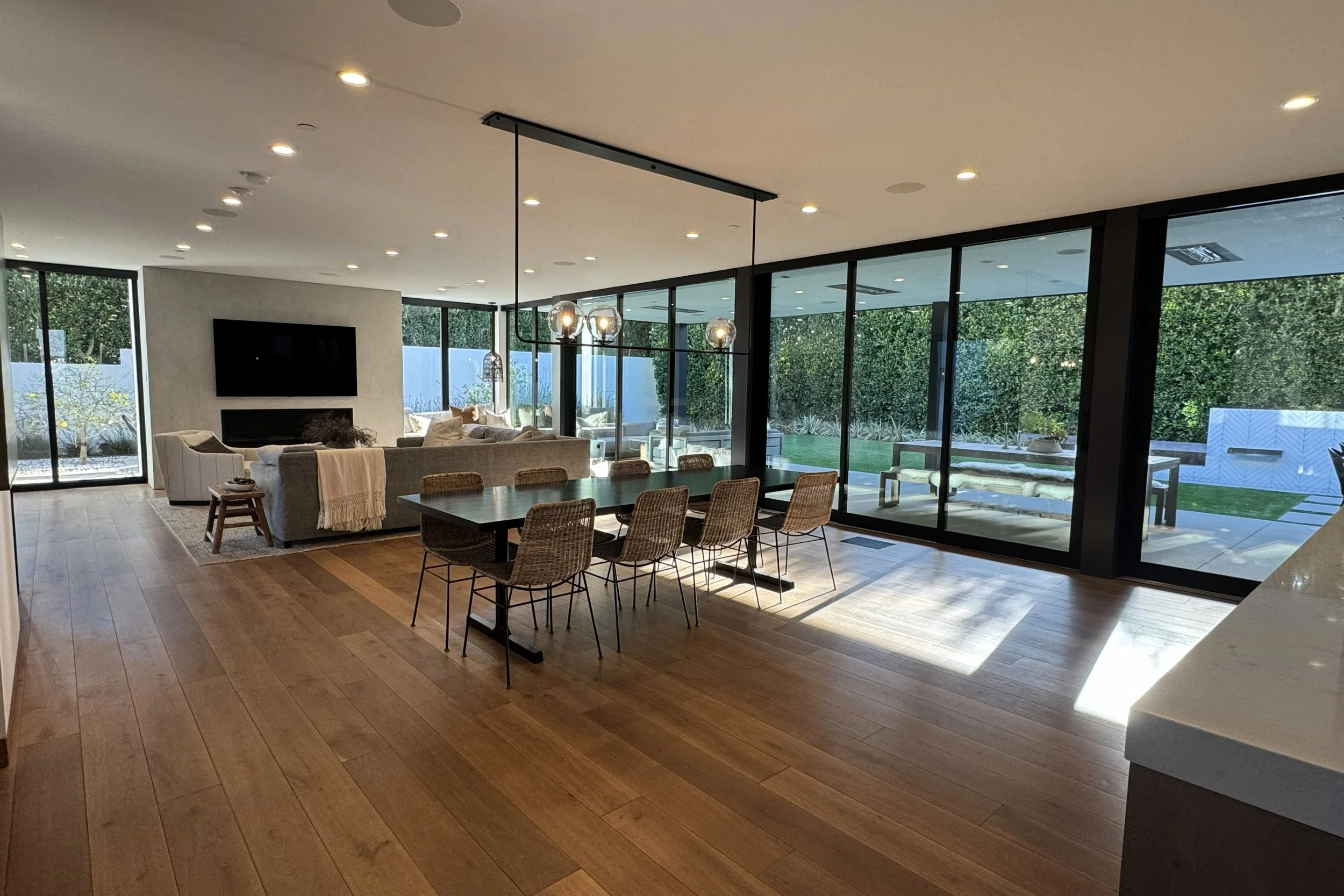 Modern open-concept living and dining area with large glass windows, wooden flooring, a wall-mounted TV, a sofa set, a dining table with chairs, and pendant lights hanging from the ceiling.