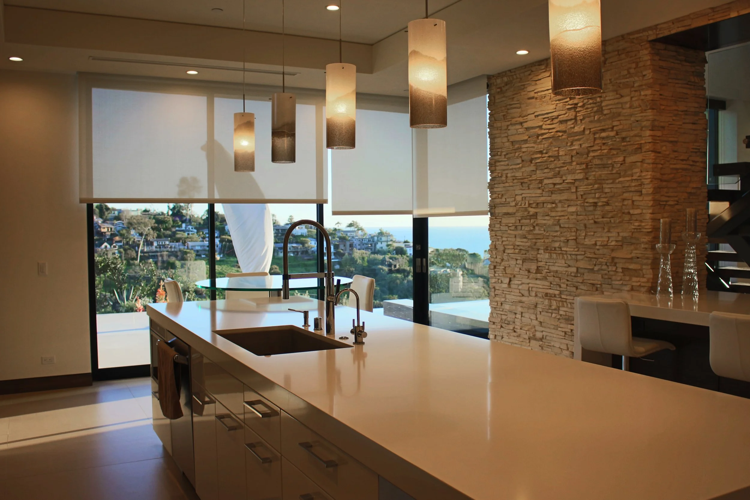 Modern kitchen with a white island, hanging pendant lights, a stone accent wall, and large windows showing a view of houses and water outside.