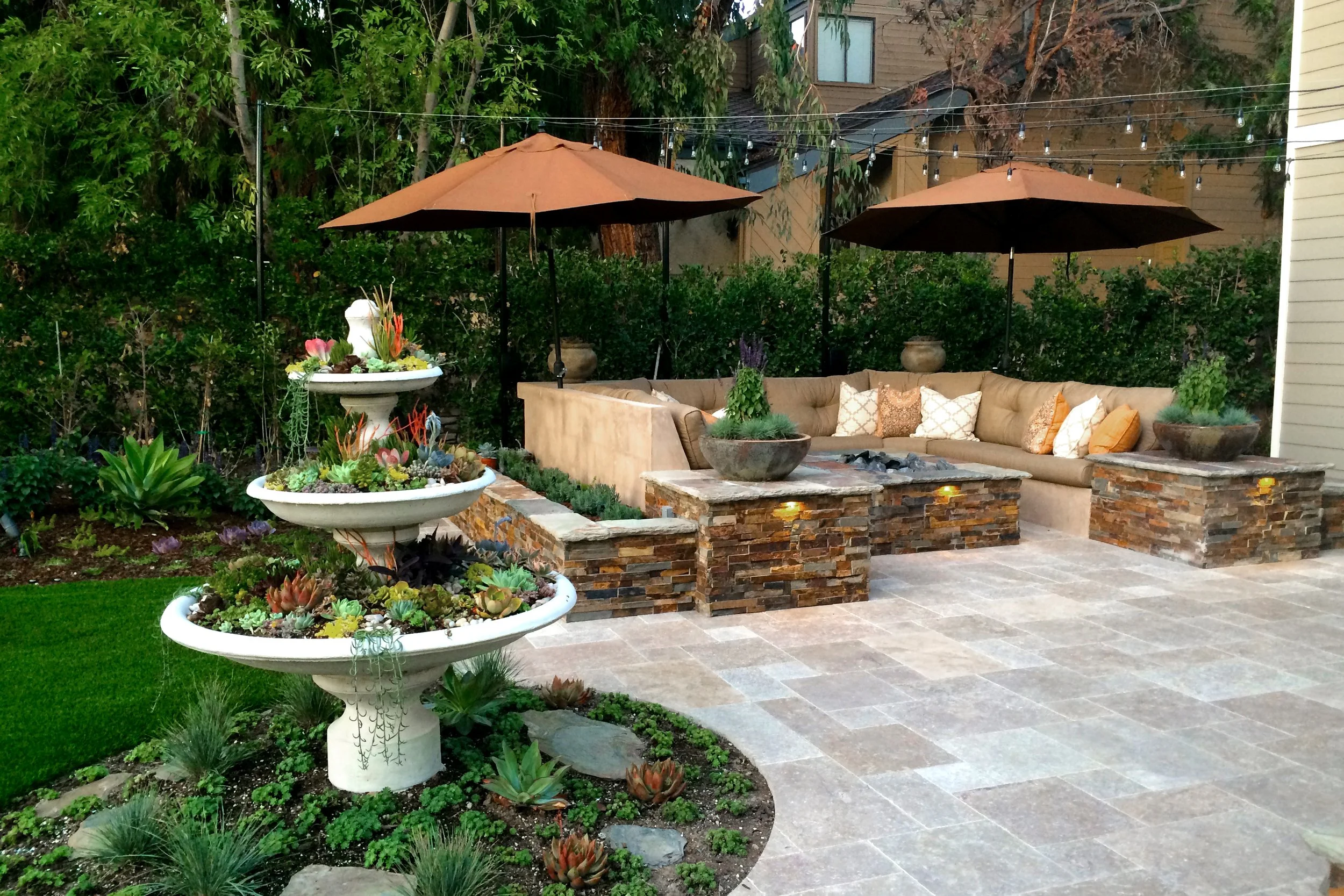 A backyard patio area with two large brown umbrellas, a curved beige outdoor sofa with decorative pillows, and a fire pit. There are potted plants, succulents, and flower arrangements in white planters, along with string lights hanging overhead. The 