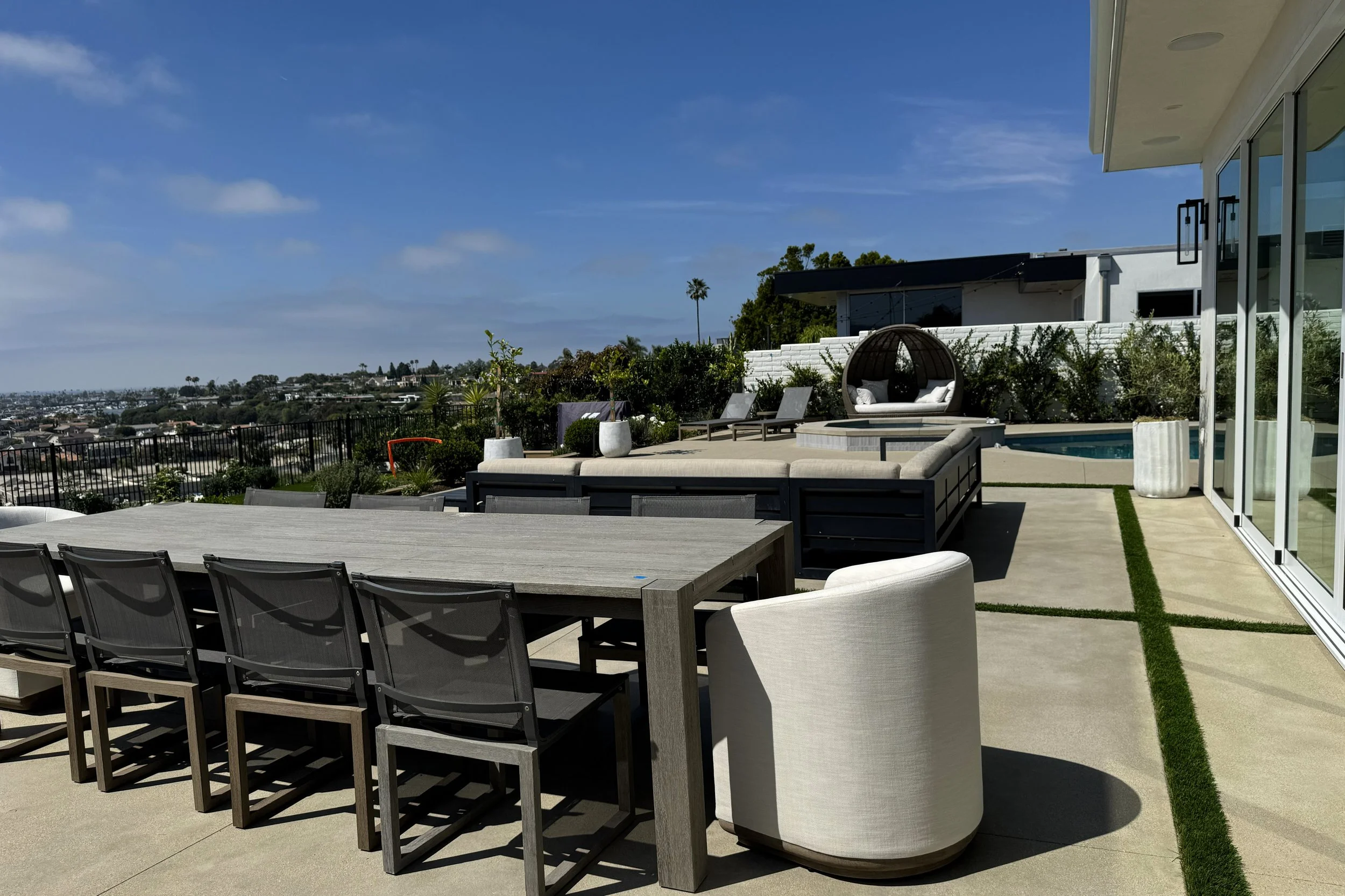Modern outdoor patio area with dining table, lounge chairs, a circular hanging daybed with cushions, and a swimming pool, overlooking a cityscape on a sunny day.