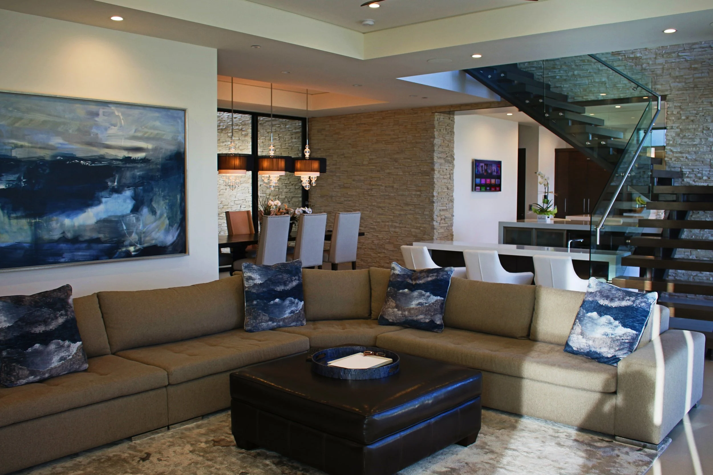Modern living room with beige sectional sofa, blue and black patterned throw pillows, black leather ottoman, abstract wall painting, dining table with chairs, stone accent wall, glass staircase, and contemporary lighting.