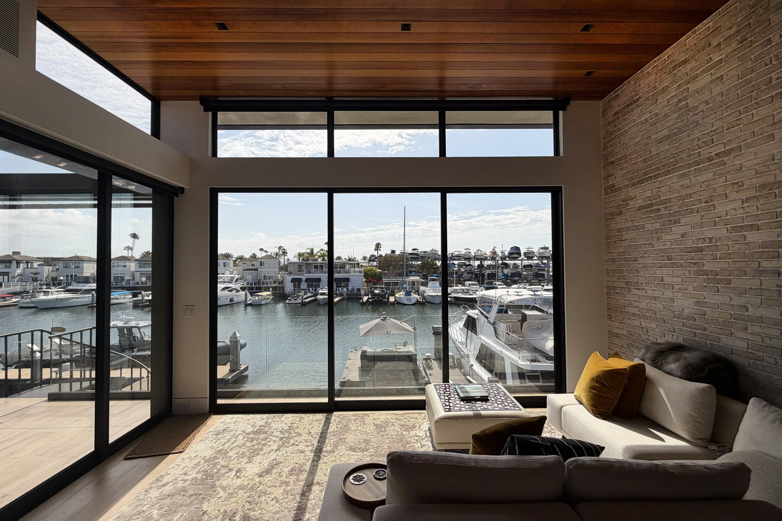 Interior of a modern house with large glass sliding doors opening to a marina full of boats, with a view of the harbor and palm trees outside.