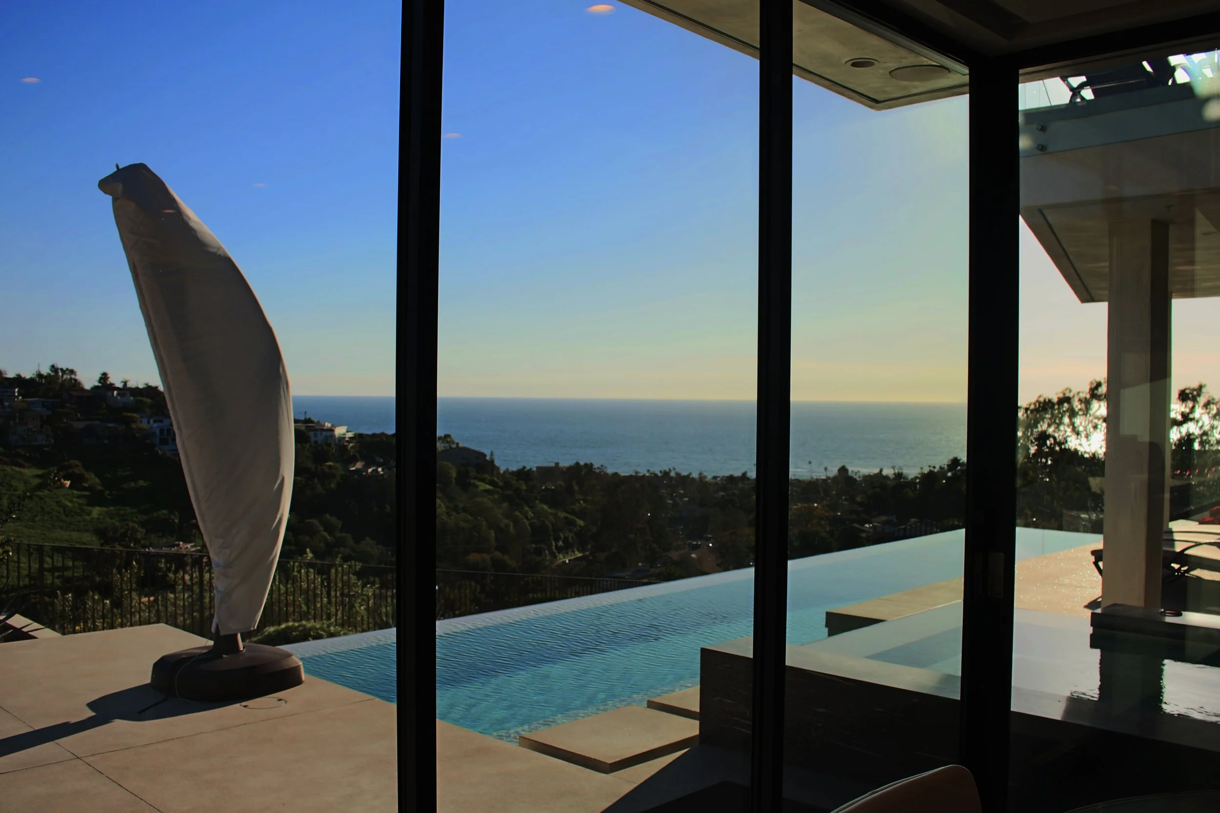 Modern living space with large floor-to-ceiling windows offering a view of the coastline and ocean. There is an umbrella on a patio beside a swimming pool and some house structures visible in the distance.