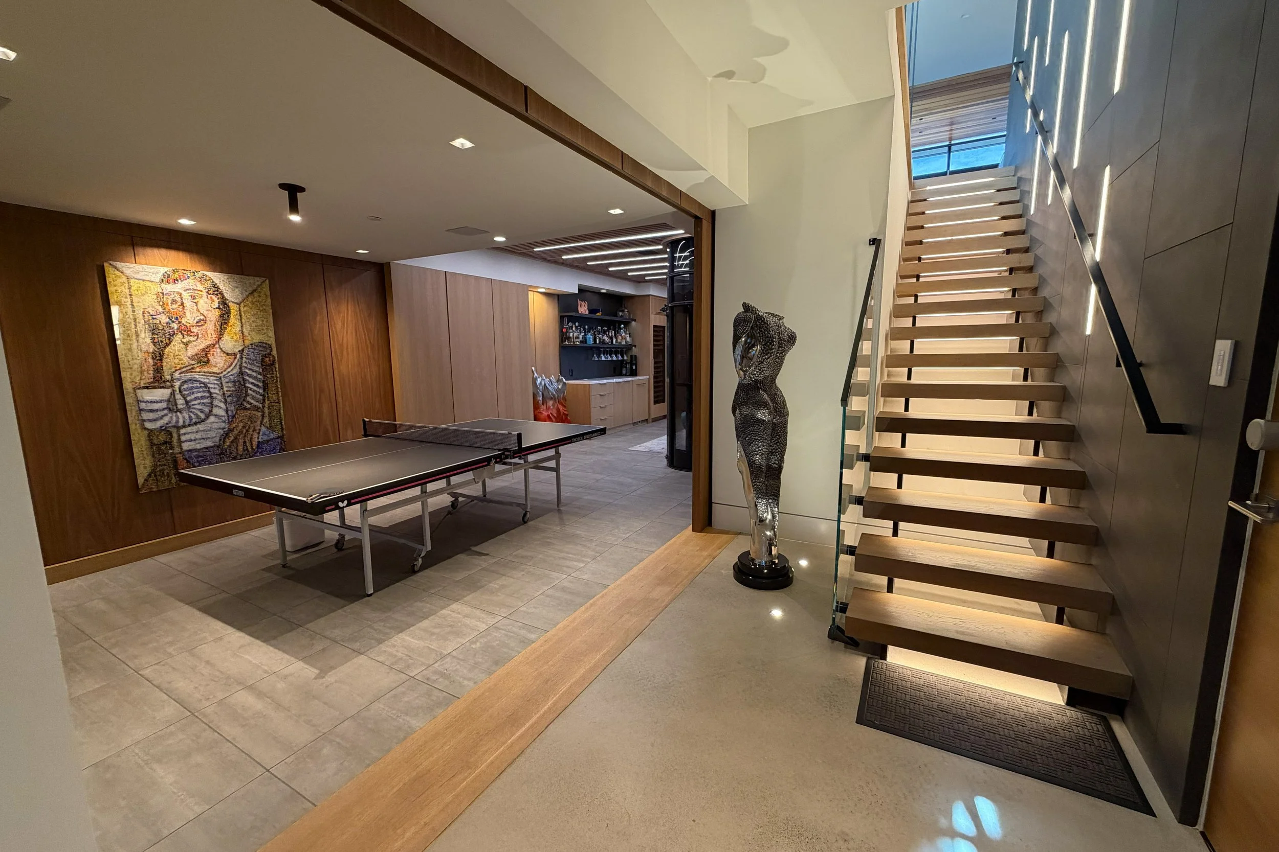 Modern stairway with wooden steps, adjacent to a sculpture of a human torso, inside a contemporary home with a bar area visible in the background.