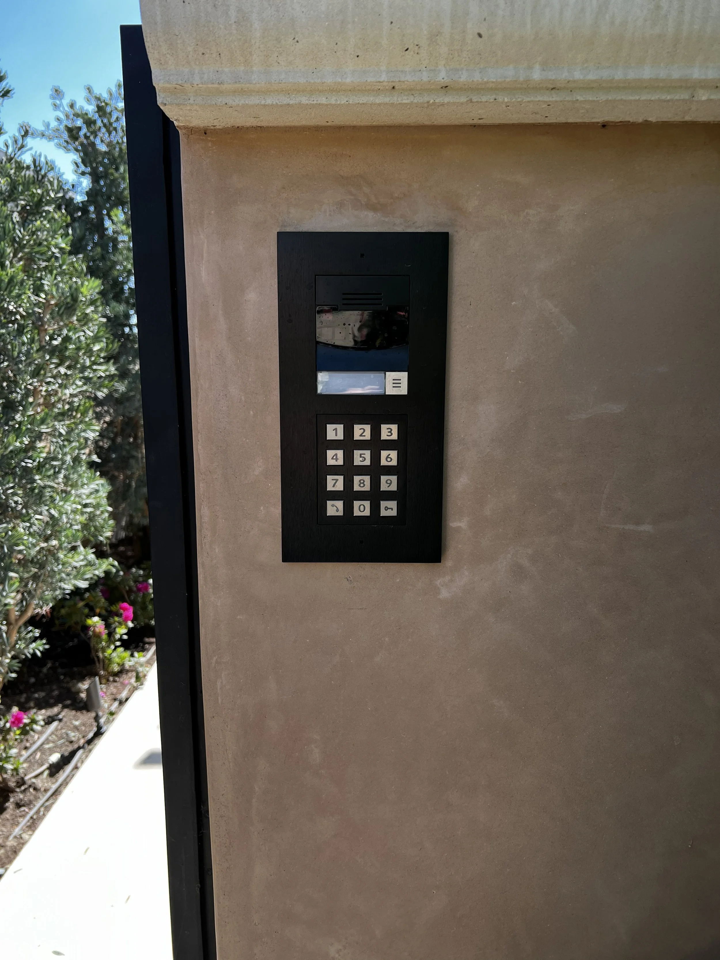 An intercom system mounted on a beige wall with a keypad and a small screen, with trees and flowers visible in the background.