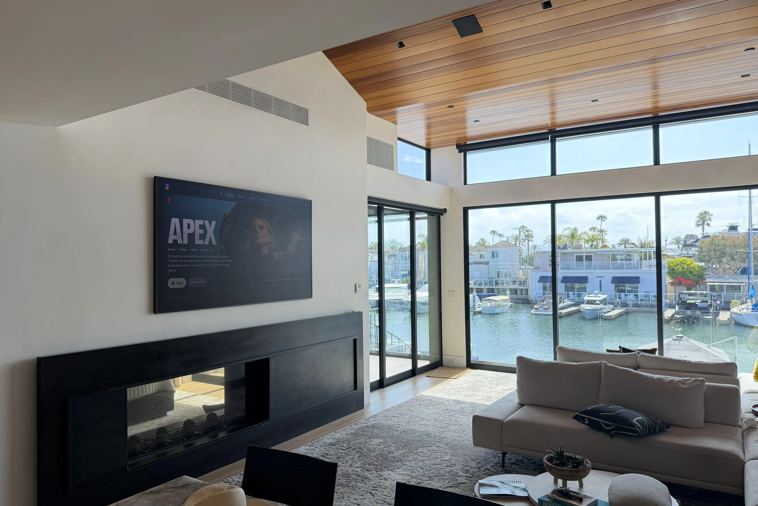Living room with a large flat-screen TV, modern fireplace, white sofa with black and white pillow, large windows showing boats and palm trees outside, wooden ceiling panels.