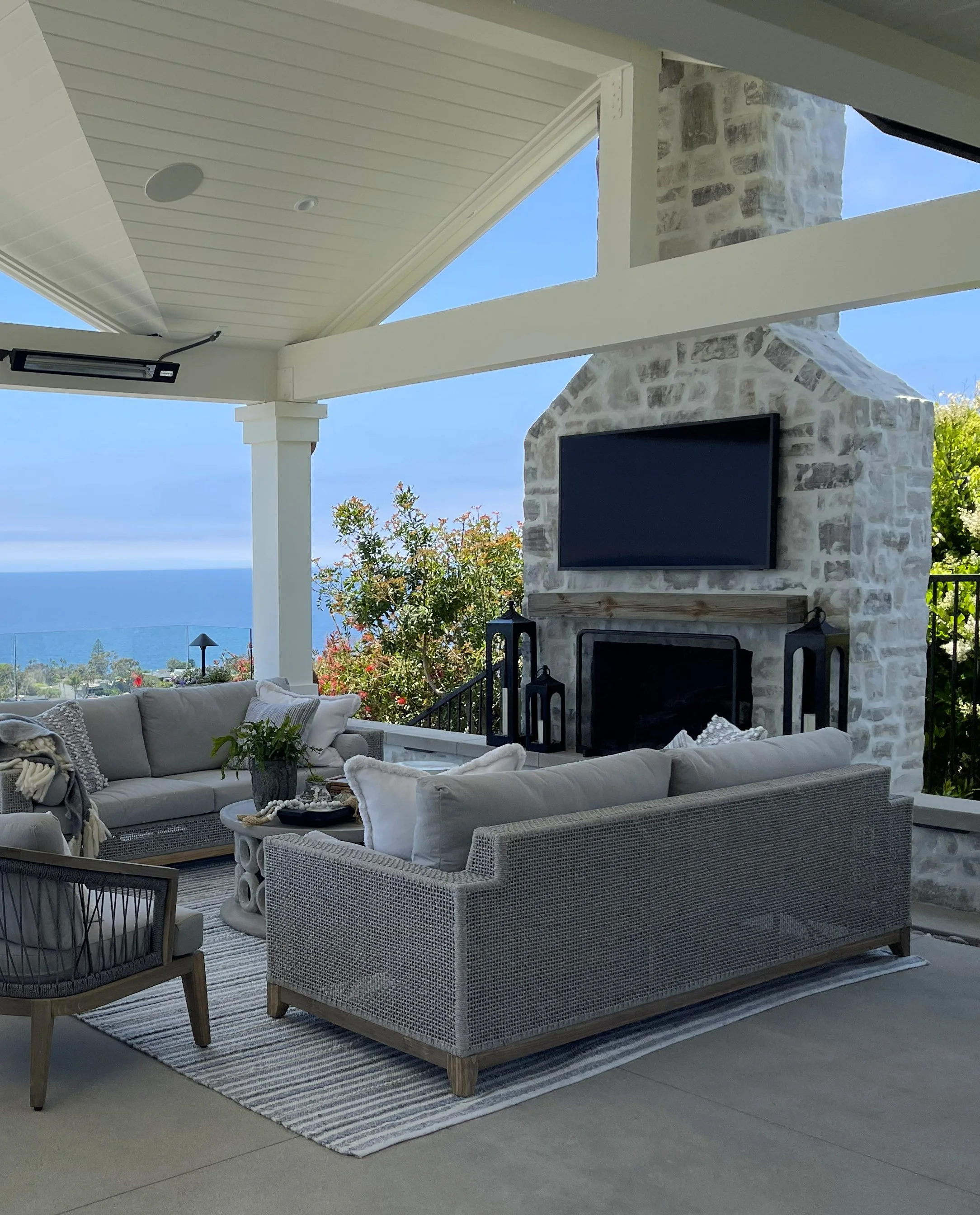 Outdoor patio with white wicker seating area, a stone fireplace with a mounted TV, plants, and a view of the blue sky and trees in the background.