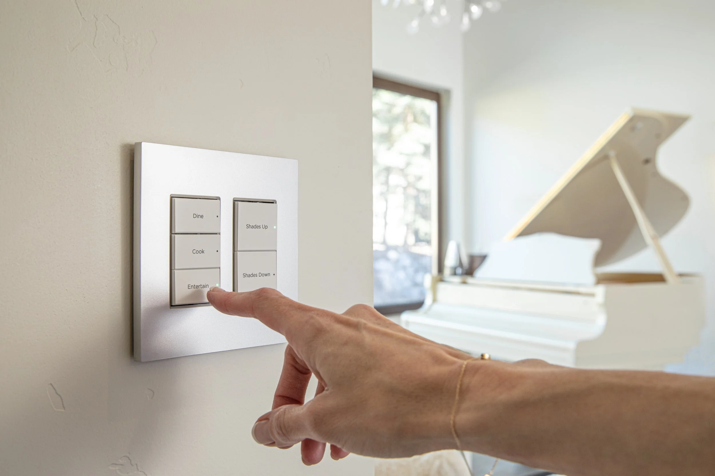 Person's hand pressing a switch on a wall-mounted control panel with labeled buttons such as 'Dine,' 'Cook,' 'Entertain,' 'Shades Up,' and 'Shades Down' in a bright room with a window and a tray table in the background.