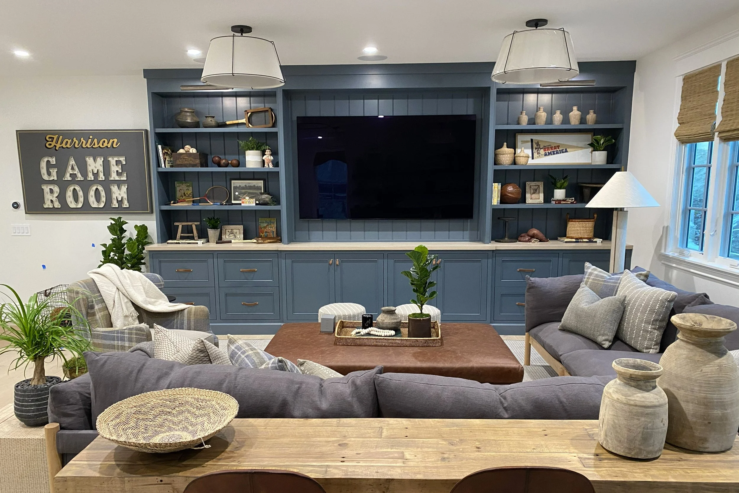 Living room with blue built-in shelves, a large TV, various decorative items, and a couch with pillows. A wooden table with vases and a tray is in the foreground. Large windows with blinds in the background.