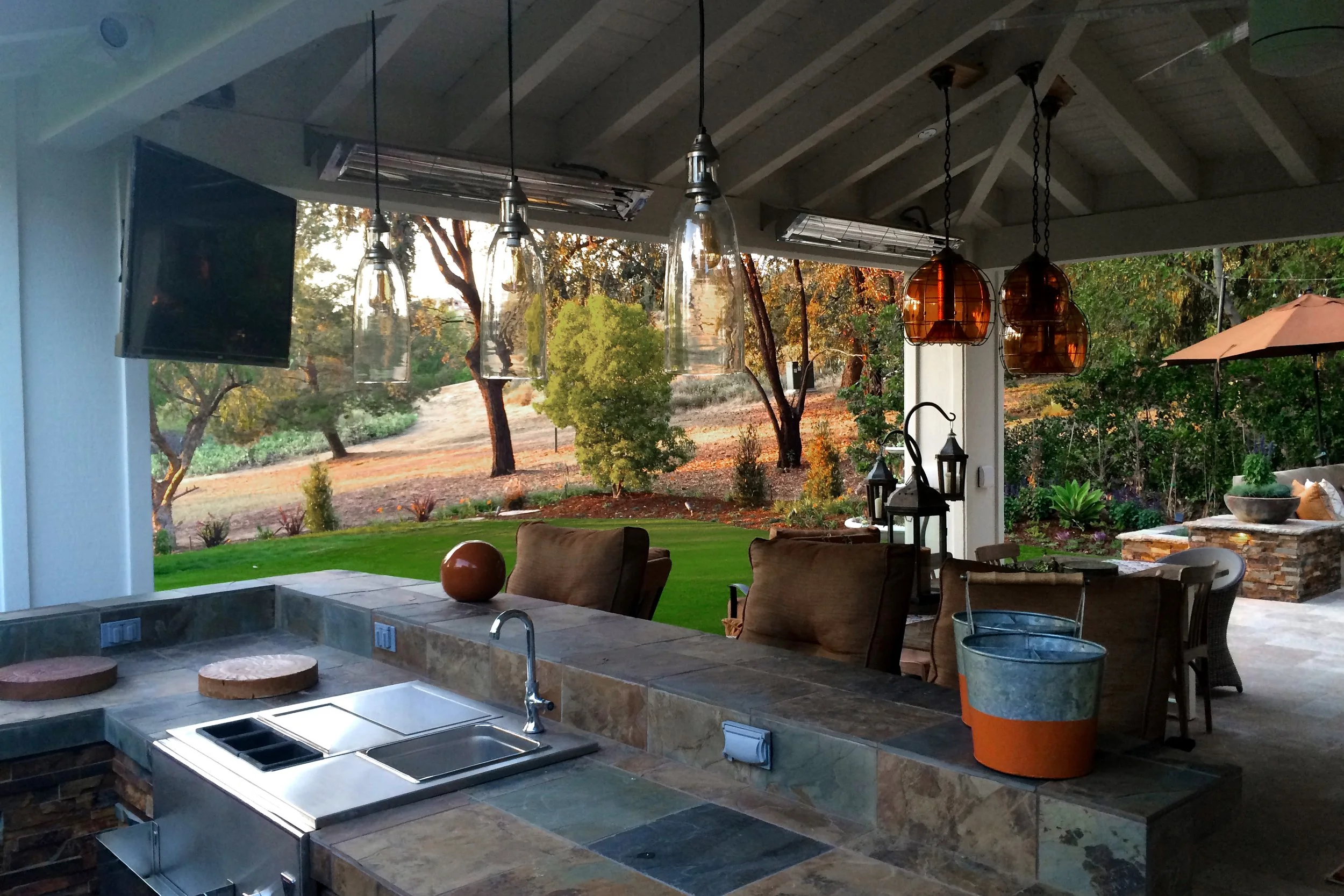 Outdoor patio with hanging pendant lights, a table with chairs, and a view of trees and a lawn in the background.