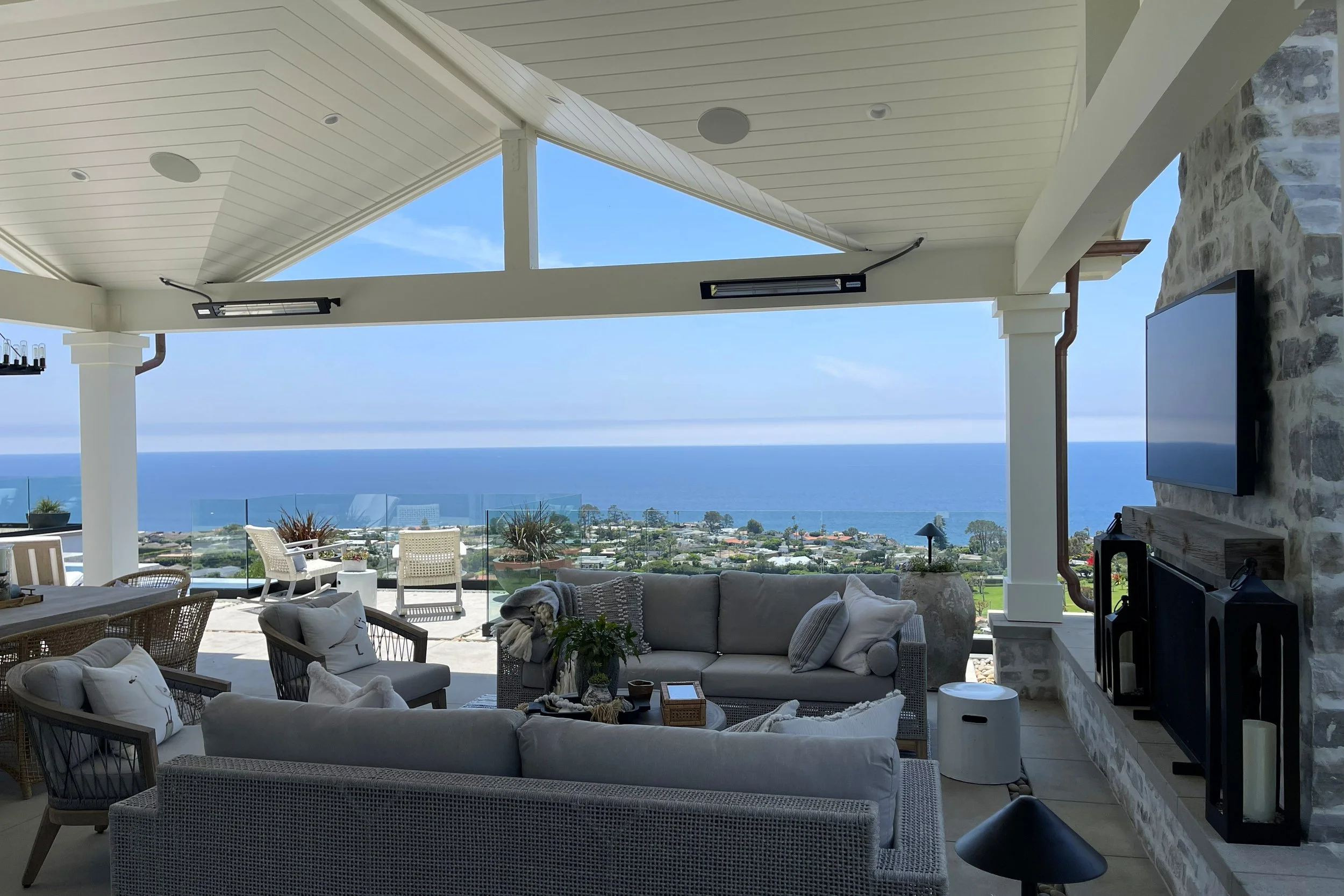 Interior of a modern living room with a view of the ocean, including white furniture, potted plants, a stone fireplace, and outdoor seating, with a partly cloudy sky.