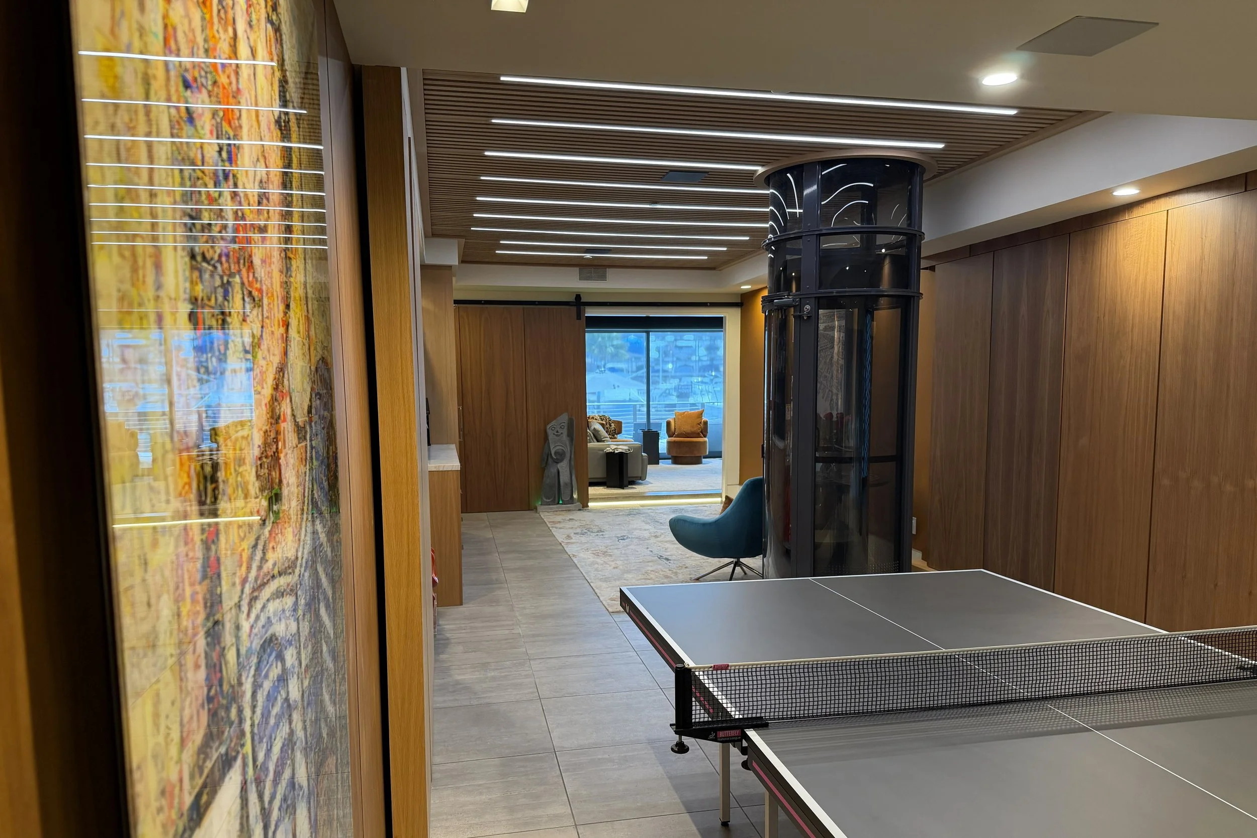 Interior of a modern recreational room with two ping pong tables, wooden wall panels, a large colorful abstract painting on the left, a black curved glass elevator or display case, a seating area with armchairs and a window showing a city view in the