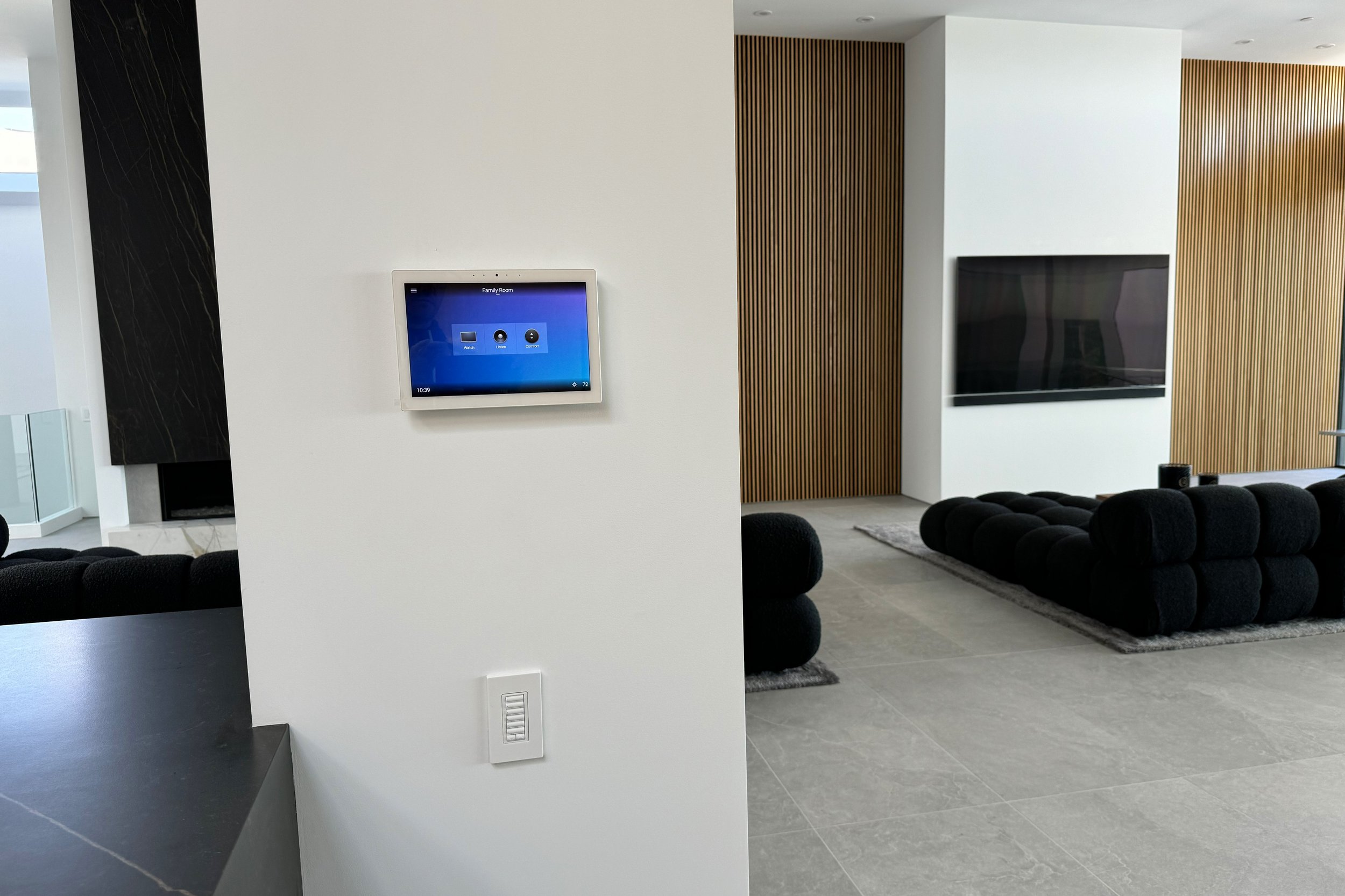 Modern living room with white walls, black tufted sofas, a wall-mounted flat-screen TV, and a wall-mounted digital control panel.