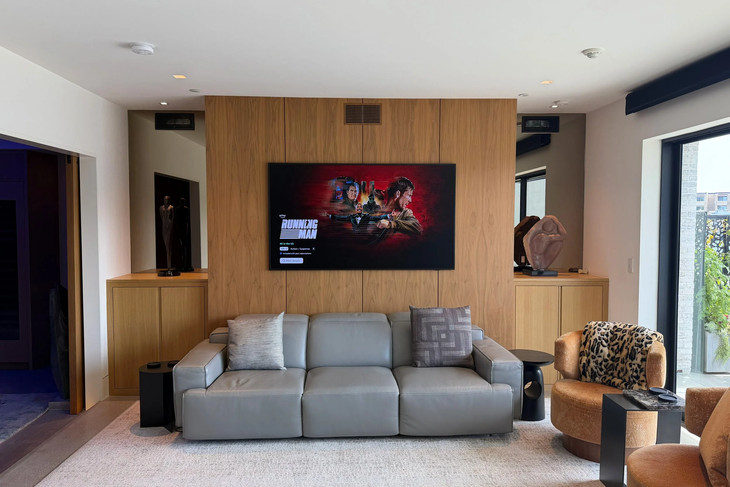 Modern living room with a light gray sofa, pillows, and a wooden wall with a large flat-screen TV displaying promotional content for the show 'Running Man.' There is a patterned armchair and a small side table on the right, with natural light coming 