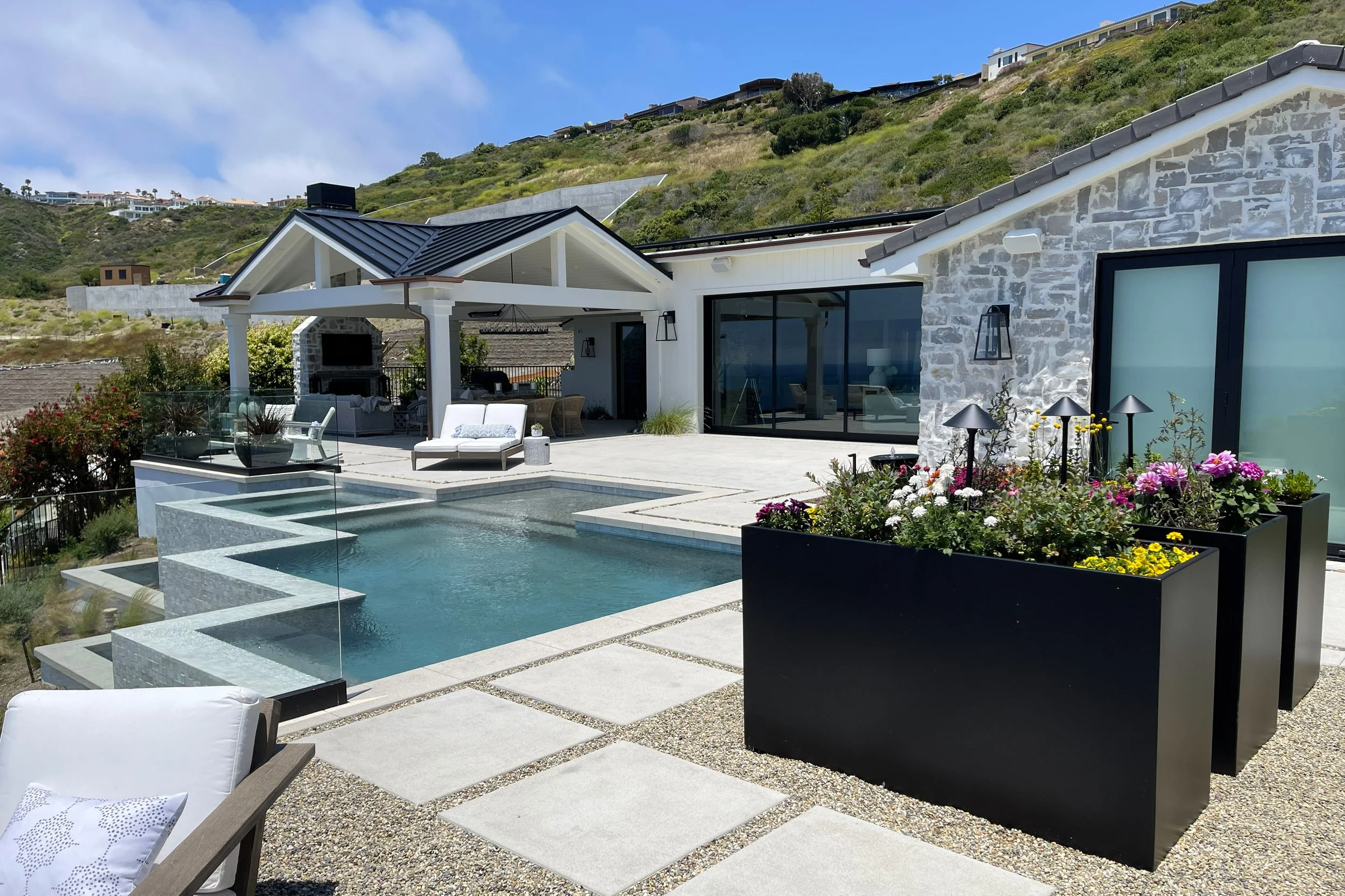 Modern house with stone facade, large black-framed windows, and outdoor living area with seating and a pool, set against a hillside with other houses on the slope.
