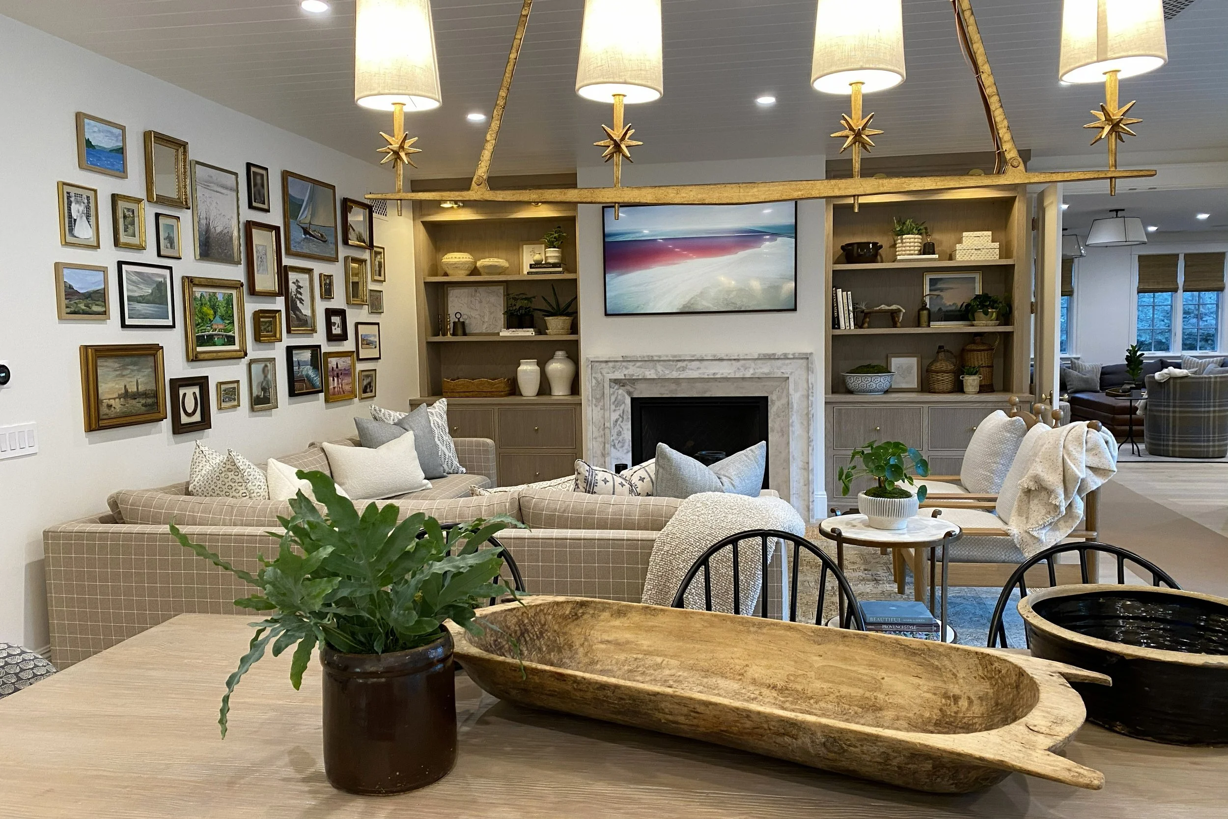 A cozy living room with beige and white furniture, framed artwork on the wall, a wall-mounted TV above a fireplace, and open shelving with decorative items. A plant is on a wooden table in the foreground. There are multiple ceiling lights, and the ro