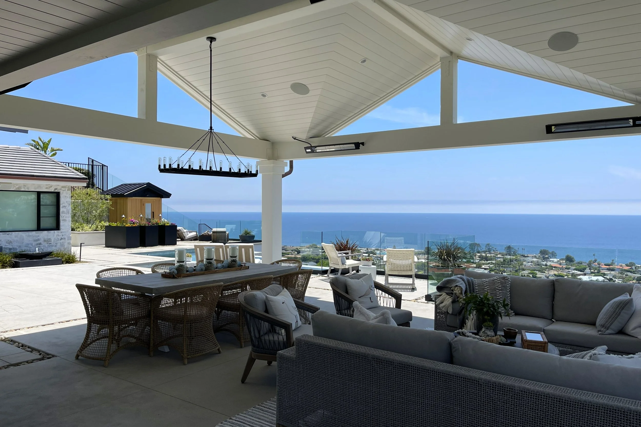 Outdoor patio area with seating, dining table, and view of the ocean under a covered roof with large windows and a chandelier.