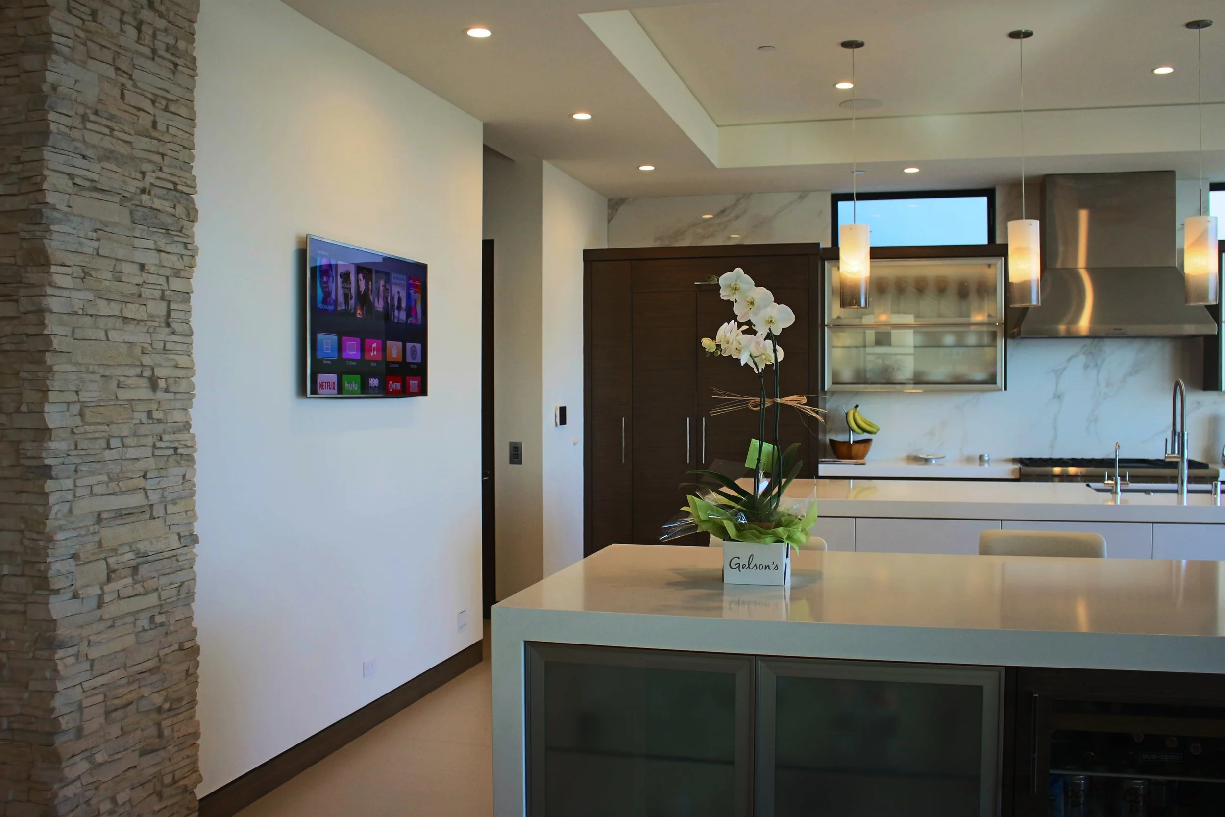 Modern kitchen with white countertops, wooden cabinets, hanging pendant lights, and a wall-mounted TV. There is a potted orchid and a bunch of bananas on the kitchen island.