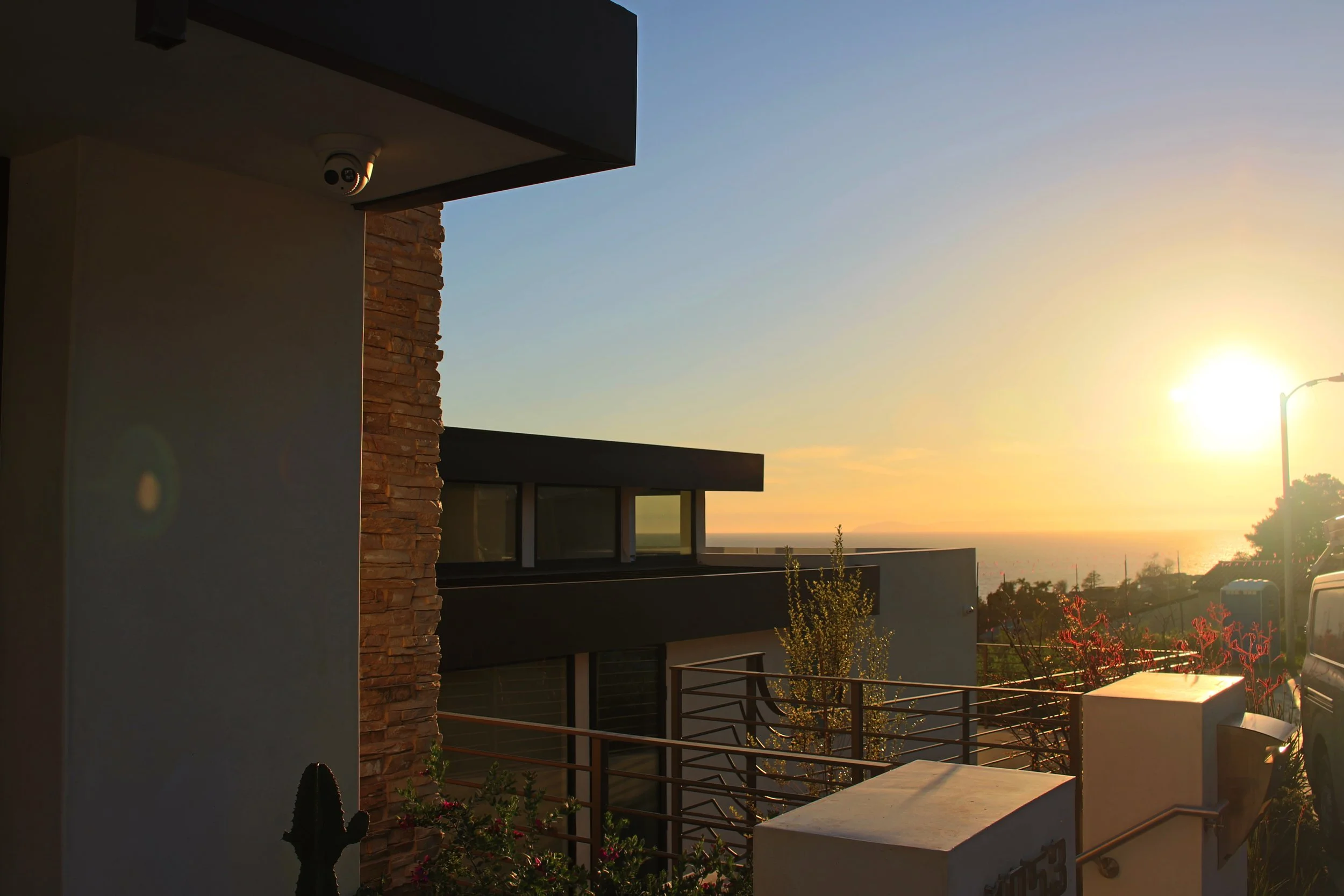 A view of a modern house exterior during sunset, featuring a cleft stone wall, a window, a rooftop with a safety camera, and a distant ocean with a brightly shining sun.