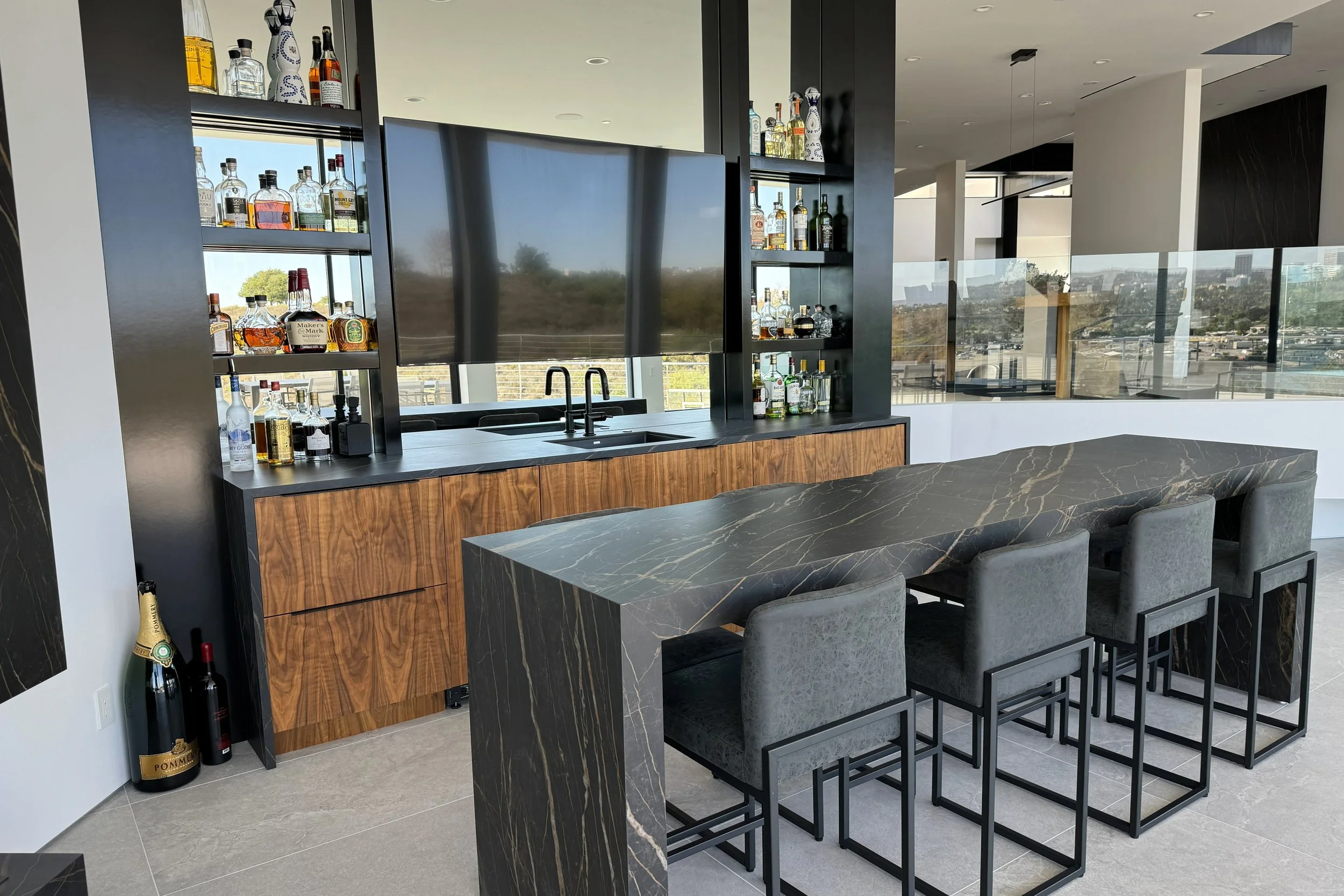 Modern indoor bar area with shelves of various liquor bottles, a large mirror, and a city view through large windows.