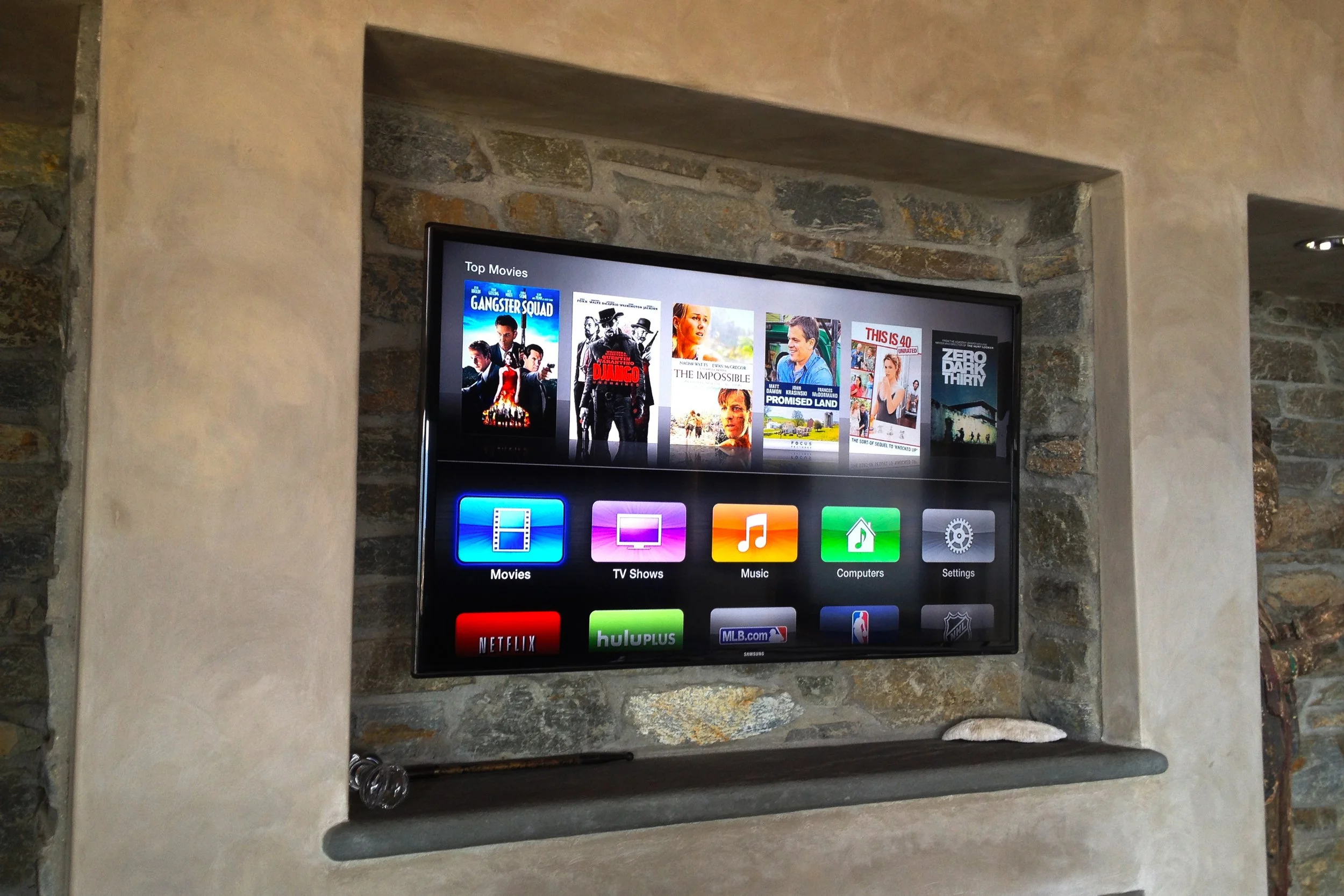 A flat-screen TV mounted on a stone wall. The TV screen displays categories including Movies, TV Shows, Music, Computers, and Settings, with several movie posters at the top.