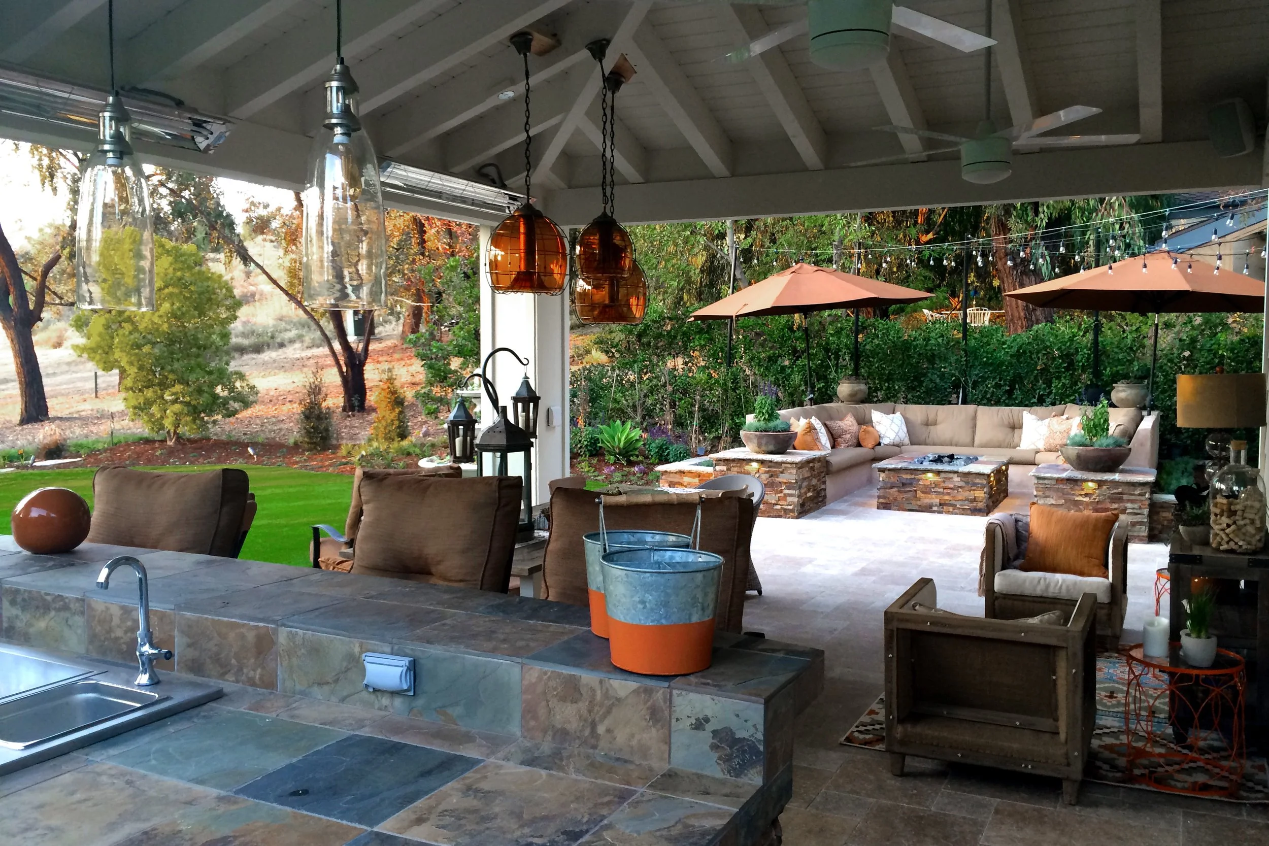 An outdoor patio area with a covered roof, hanging pendant lights, sitting area with a sectional sofa under umbrellas, and a garden with trees and landscaping.
